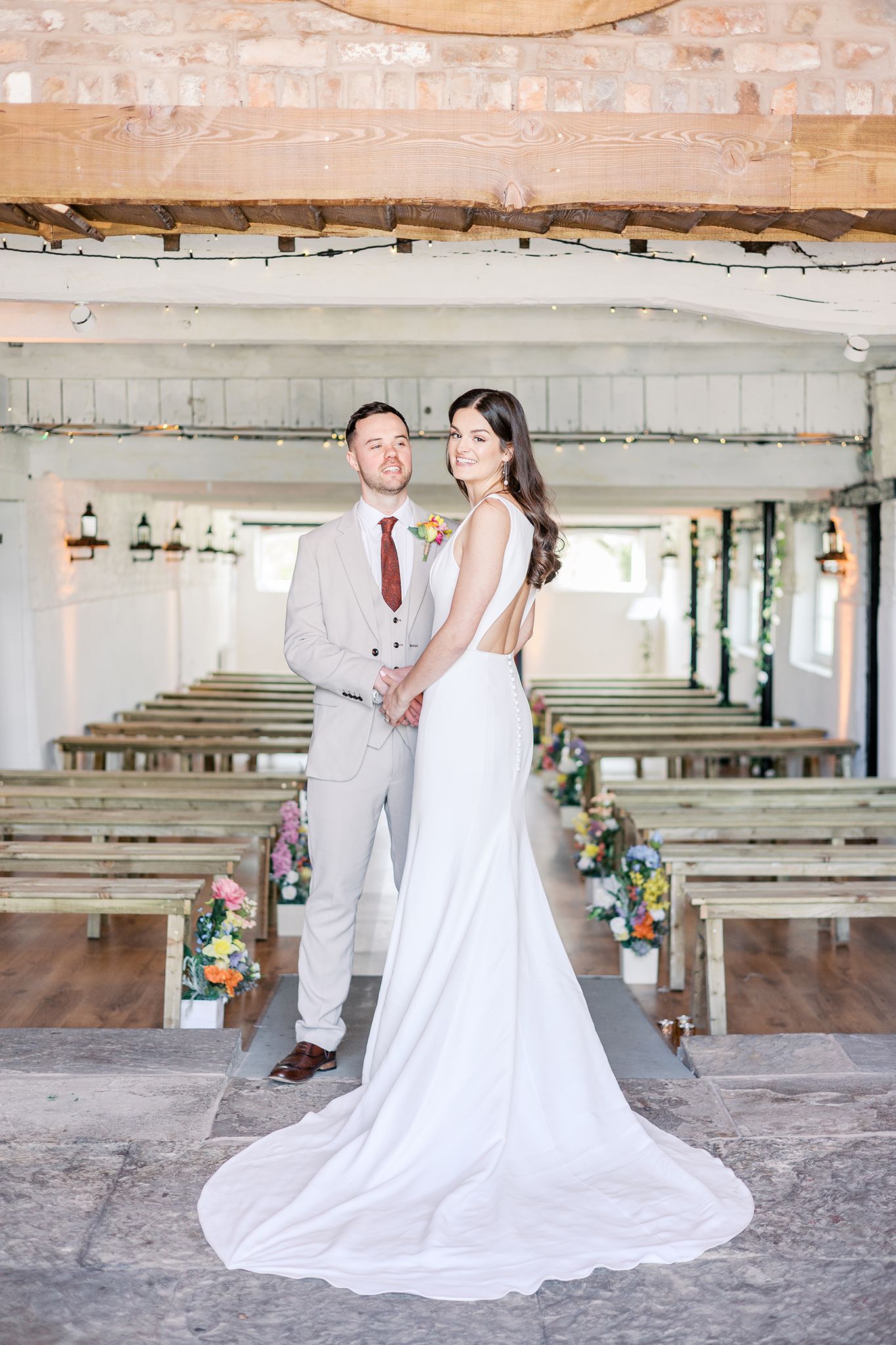 A couple stands in a rustic wedding venue with wooden church pews and floral decorations along the aisle.