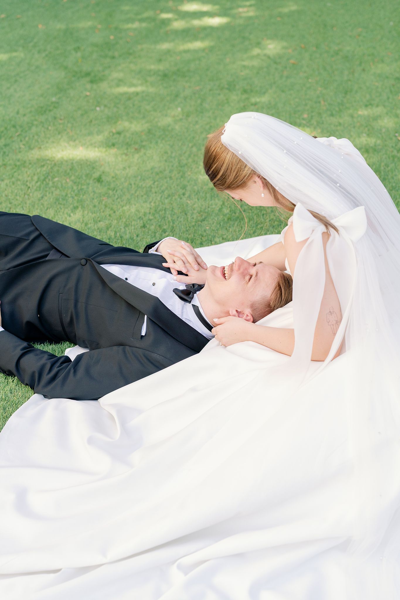Groom lies on green grass as bride in white gown and veil leans over him tenderly during wedding portraits.