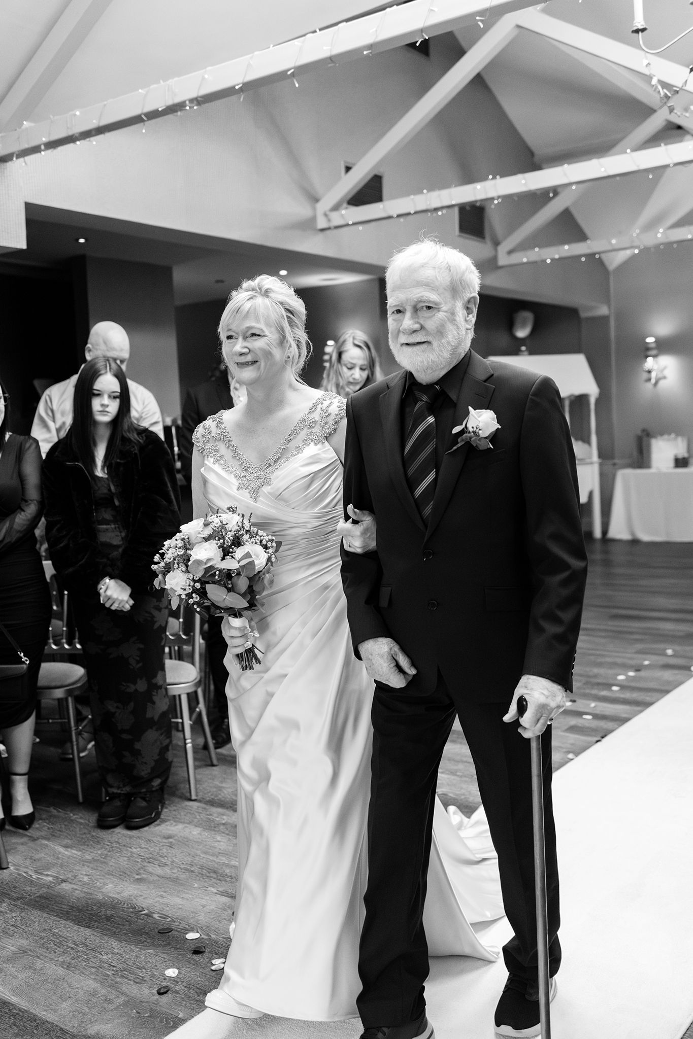Black and white photo of formal wedding processional on wooden floor with exposed ceiling beams and elegant decor.