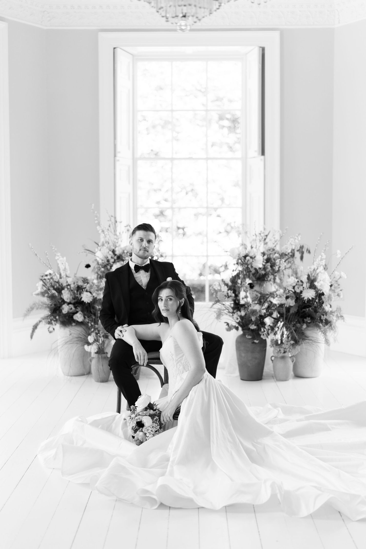 Black and white portrait of bride and groom posing elegantly in a bright room with large floral arrangements.