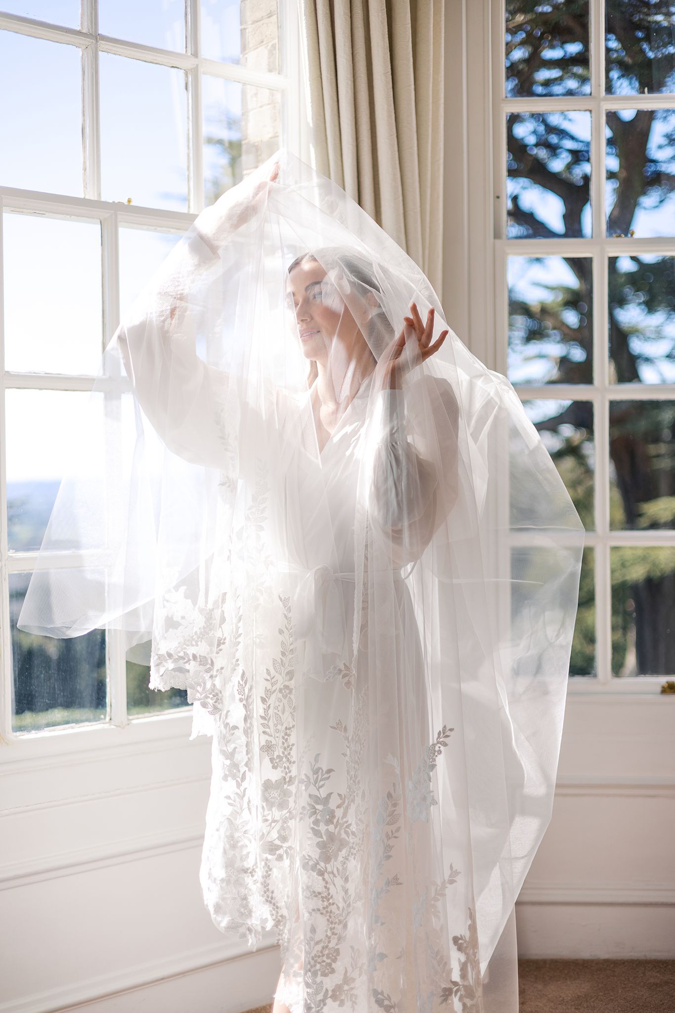 A flowing white wedding dress with delicate lace details is illuminated by sunlight streaming through large windows.
