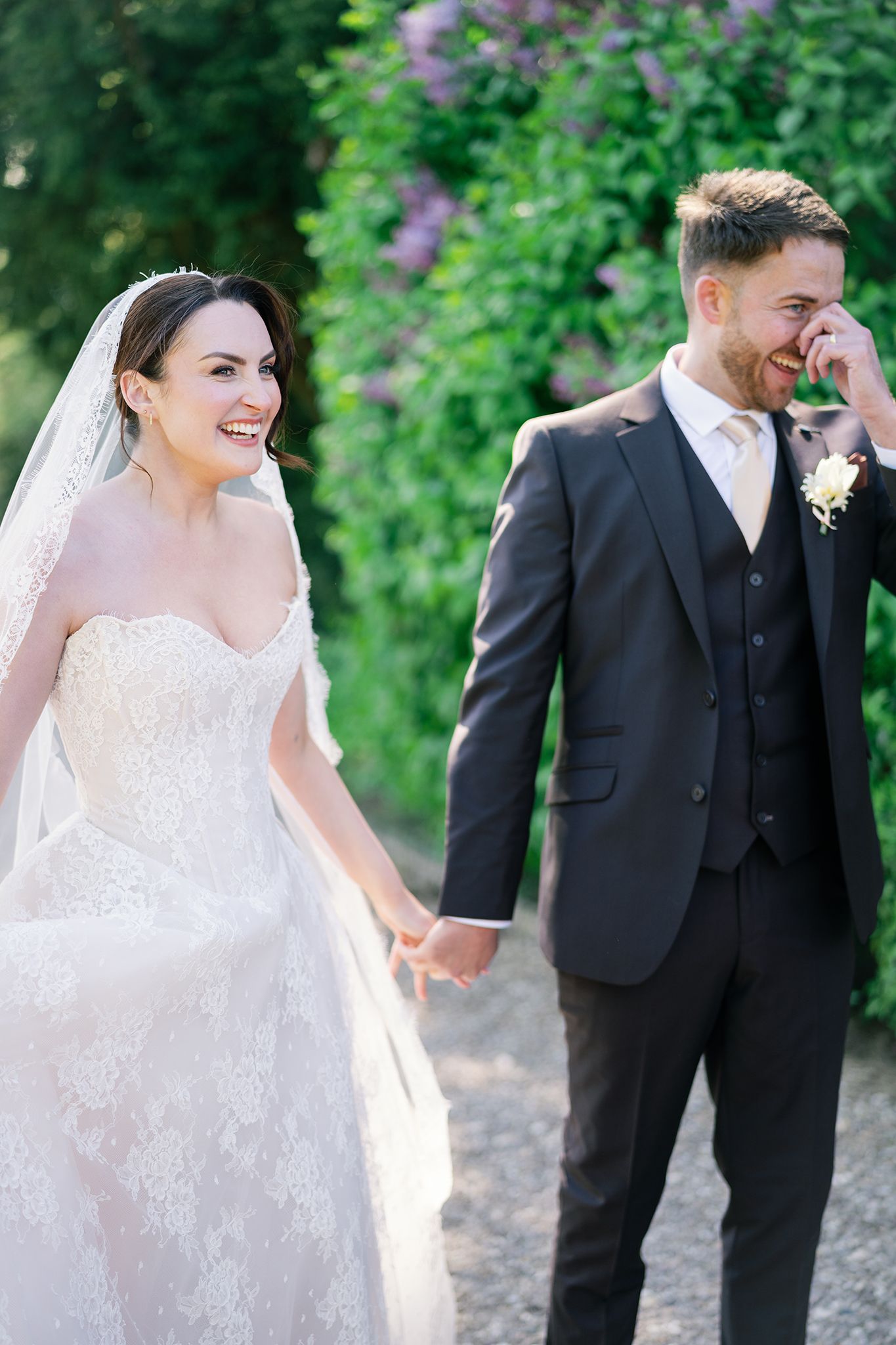 Bride and groom laughing and holding hands outdoors, bride in lace gown with veil, groom in dark suit with boutonniere.