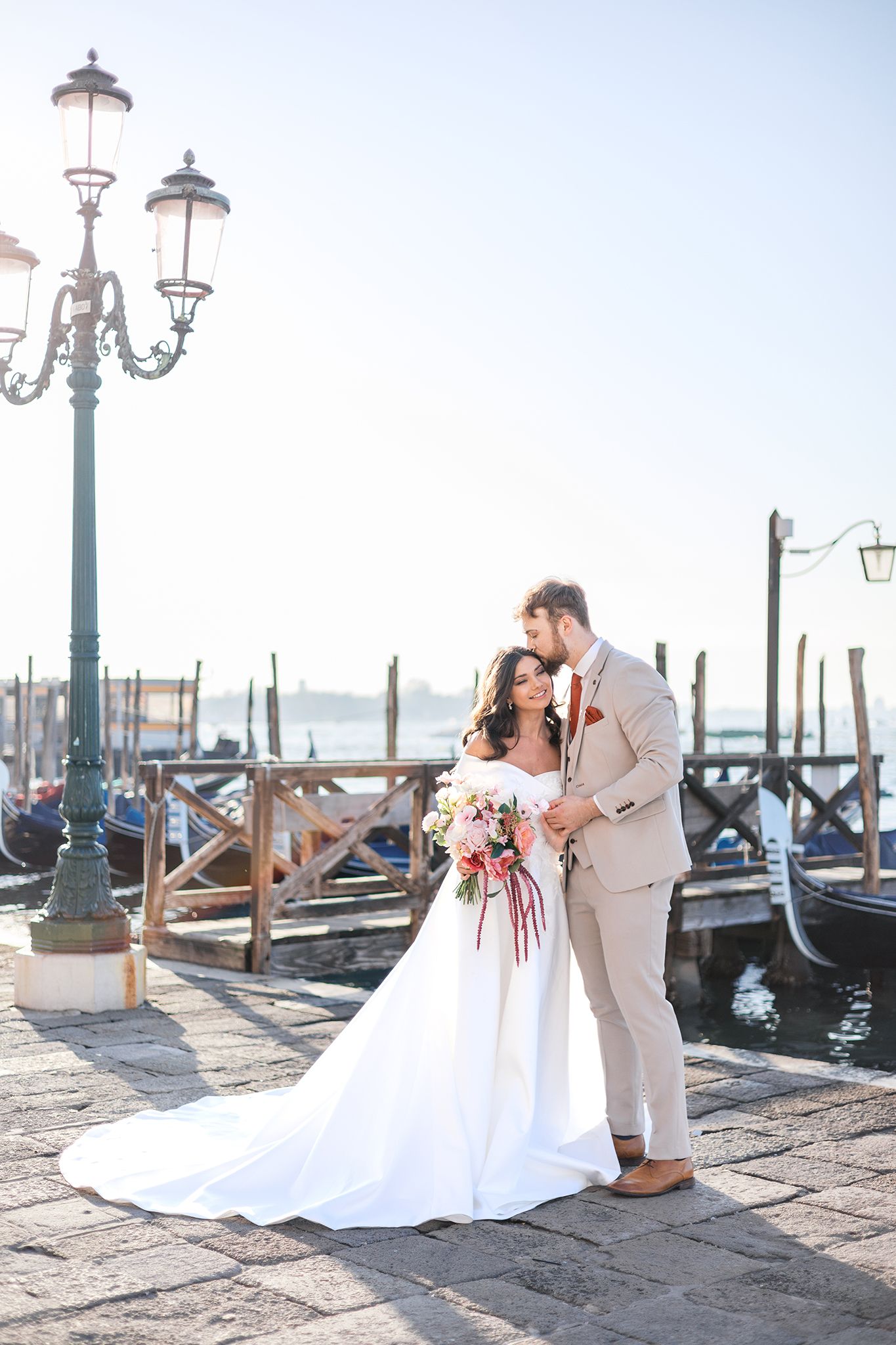 A romantic wedding couple embraces along the canal in Venice, Italy with gondolas and ornate street lamps in the background.