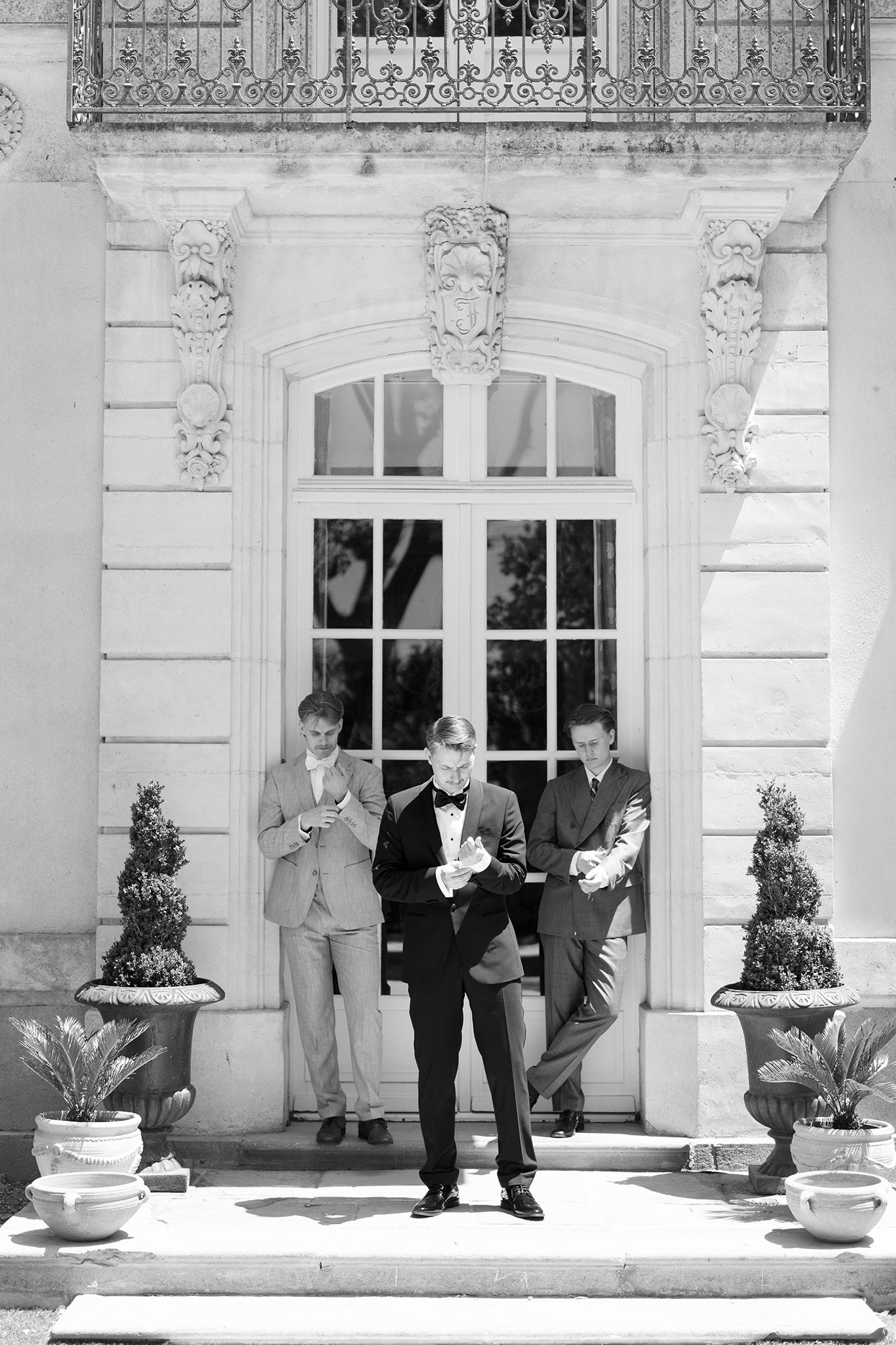 Black and white photo of three groomsmen in suits standing on elegant building steps with potted topiaries.
