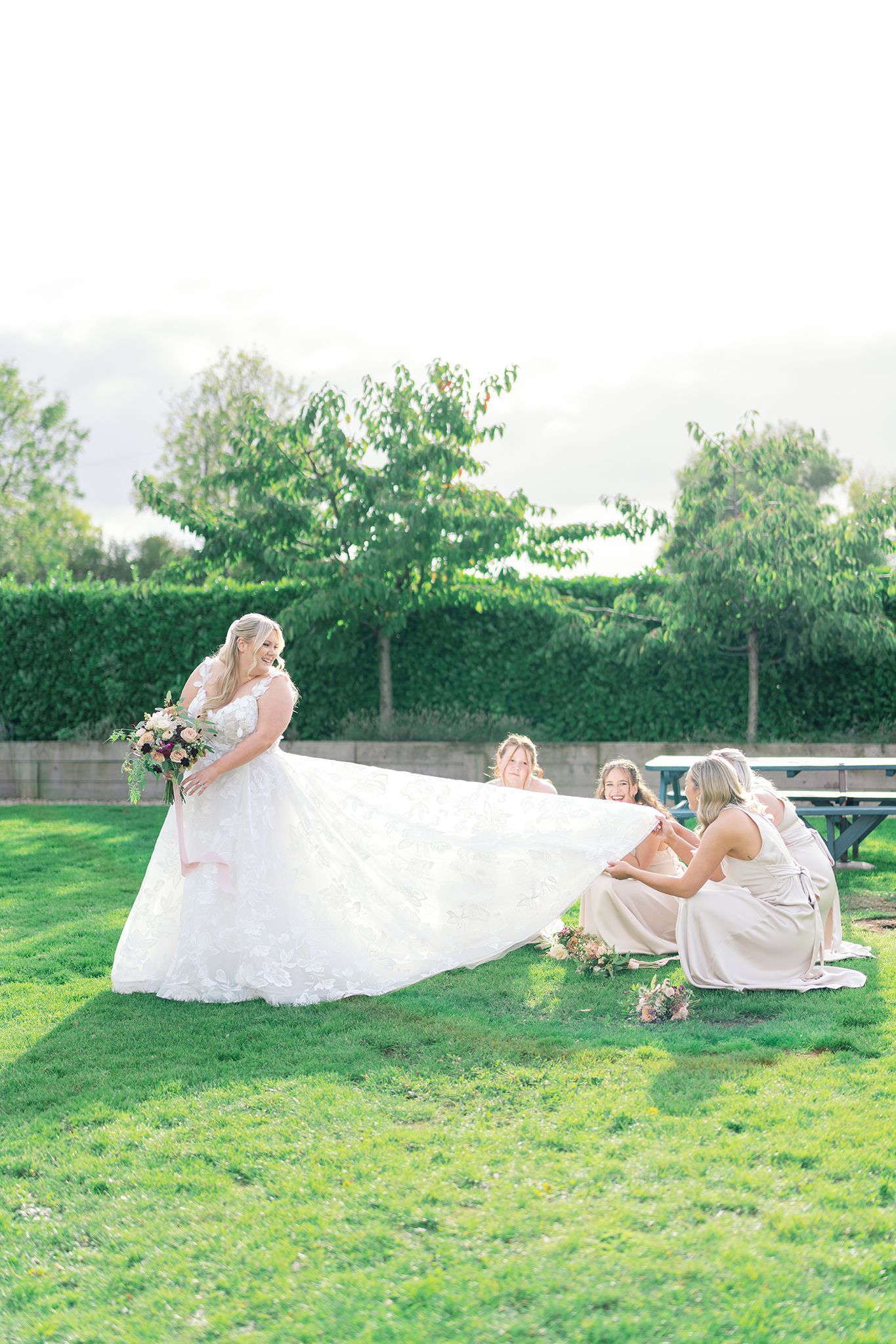 Bride in a flowing white lace gown holds a bouquet while bridesmaids in champagne dresses spread her long train on a sunny green lawn.