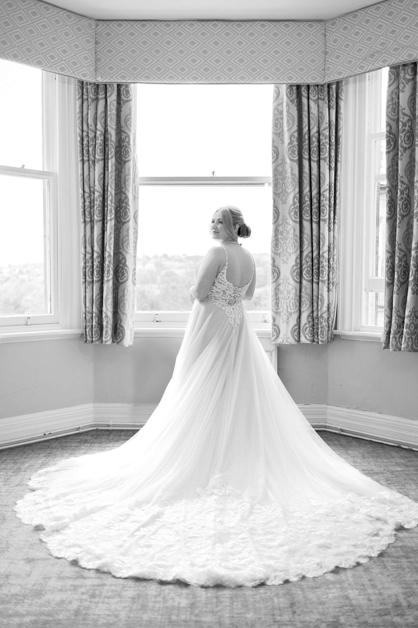 A bride in a flowing white gown with lace detail and long train stands elegantly by bay windows in a vintage room.
