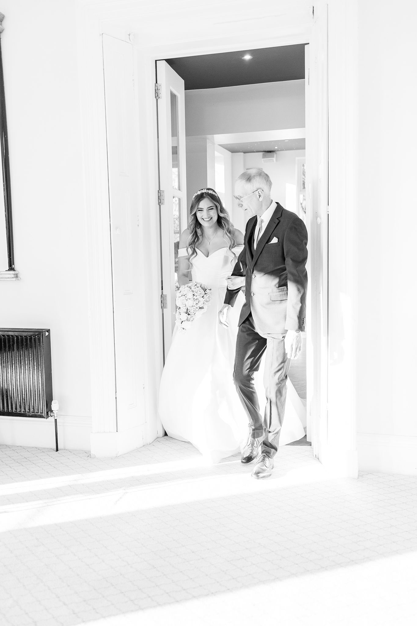 Black and white photo showing a bride in a white gown and a distinguished companion entering through a doorway.