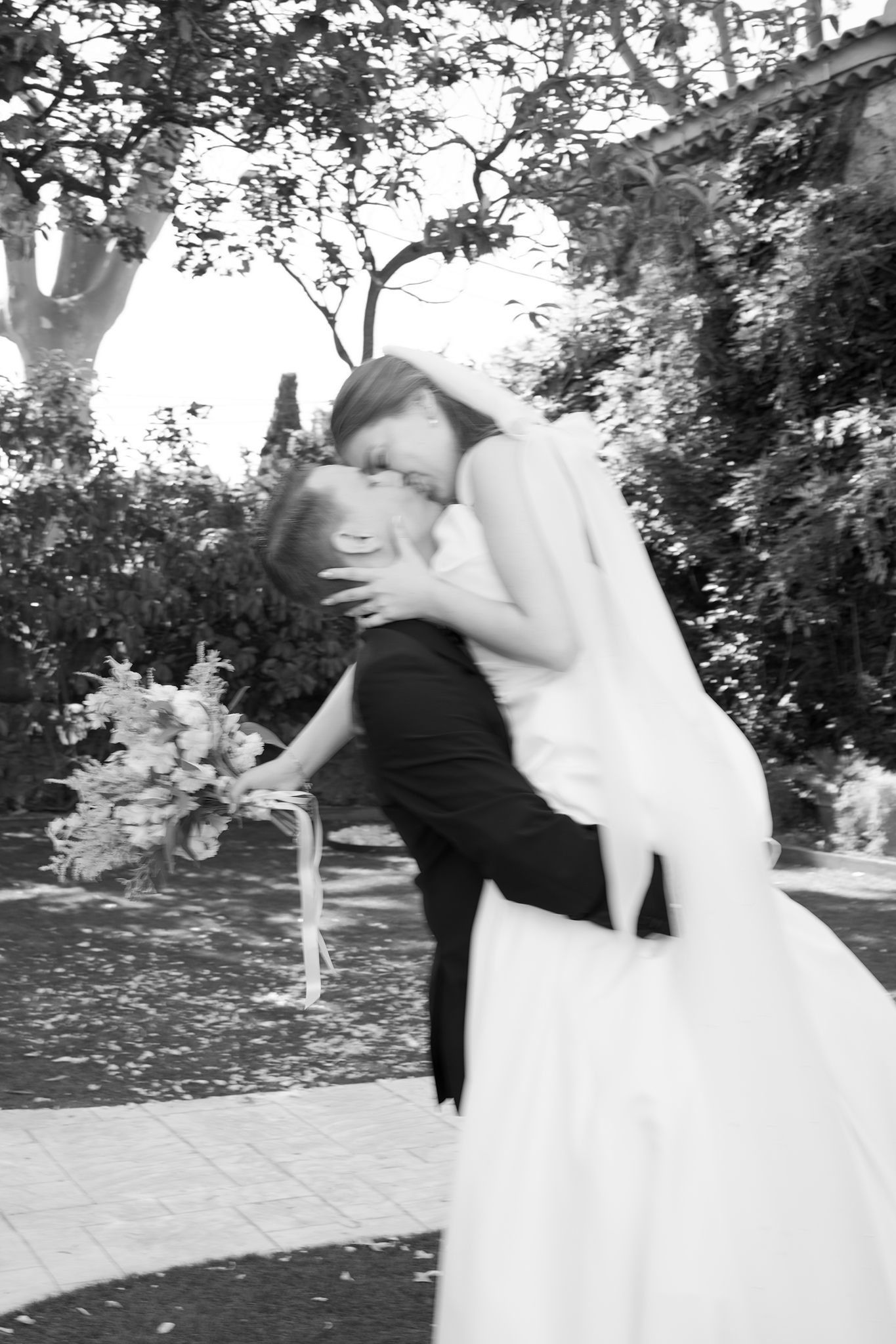 Black and white photo of a bride and groom sharing a romantic dip kiss outdoors, bride holding a bouquet.