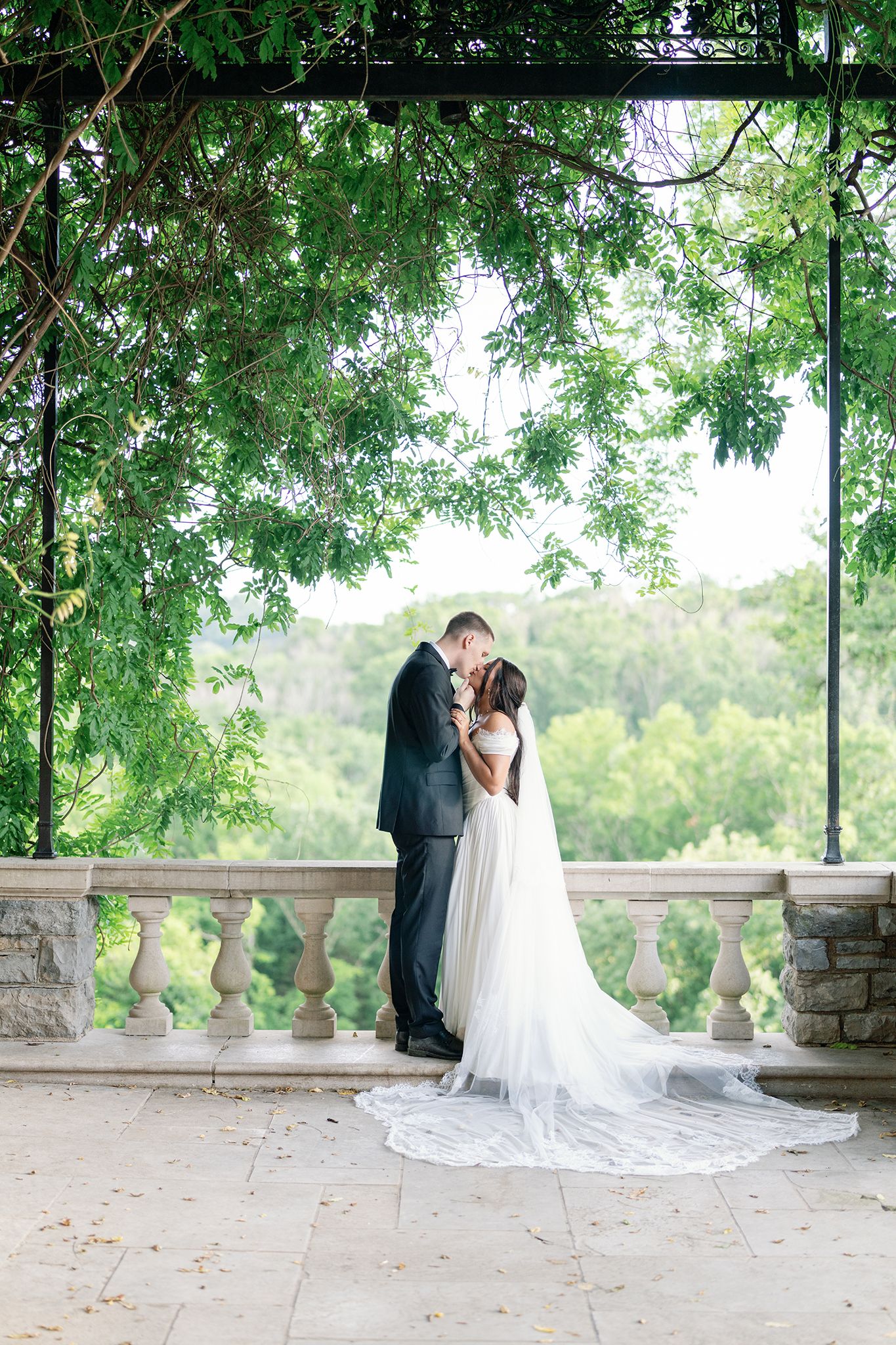 Bride and groom sharing a romantic kiss under a lush green pergola on a stone balcony in an elegant outdoor wedding setting.