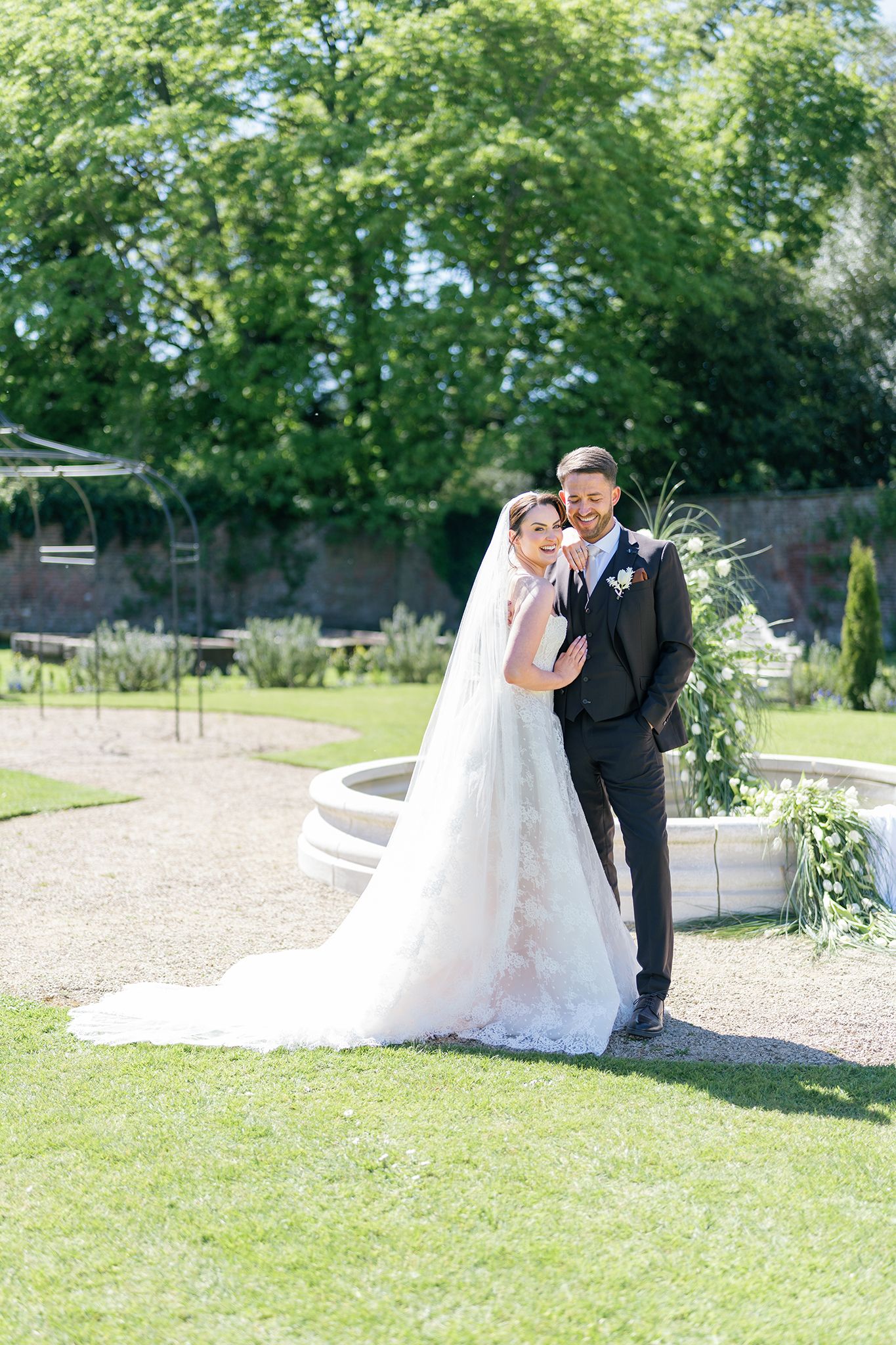 Bride in white lace gown and veil poses with groom in black suit in a lush garden with fountain backdrop.
