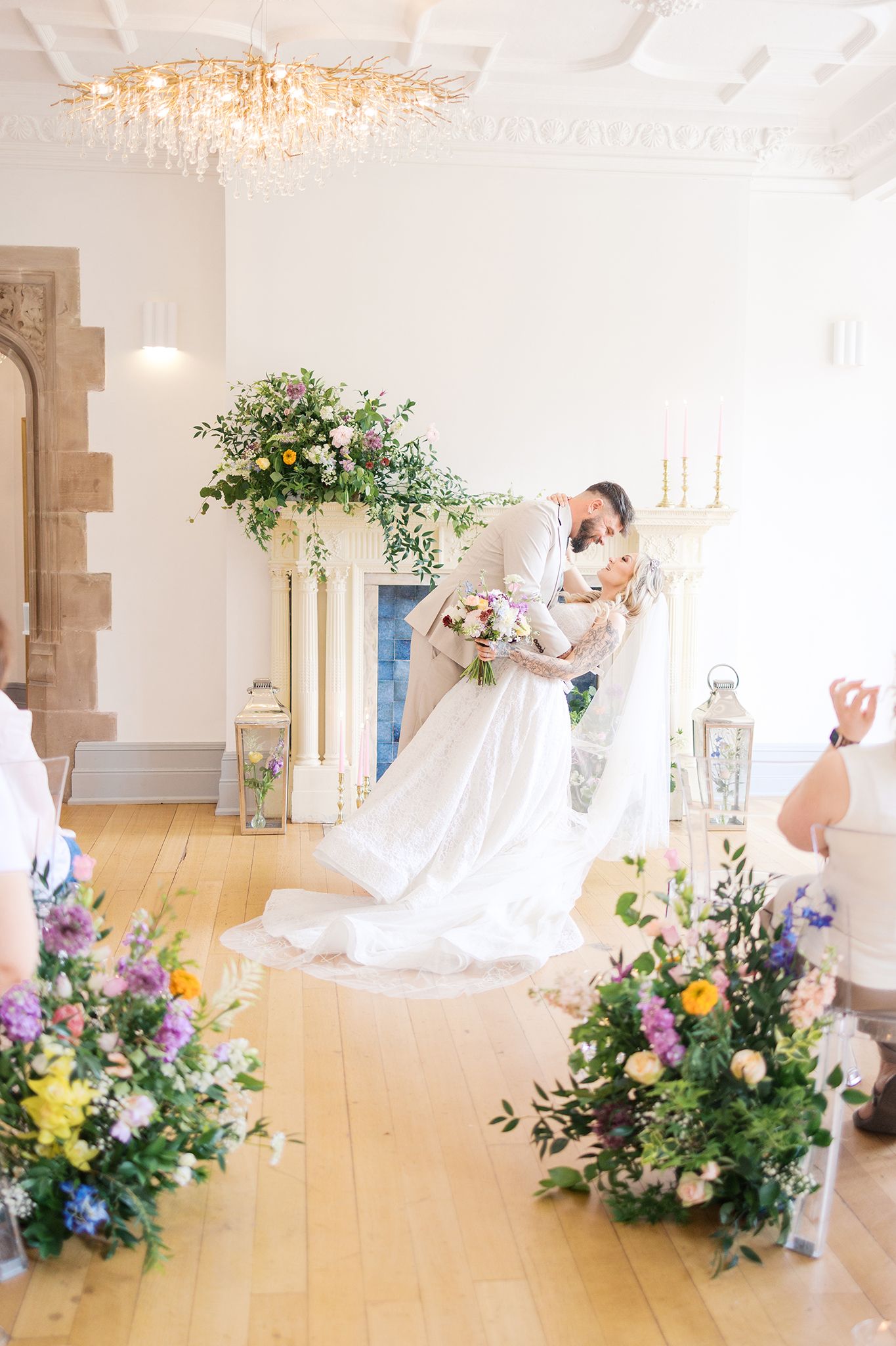 Romantic wedding scene in light-filled ballroom with elegant spring floral displays on hardwood floors beneath crystal chandelier.