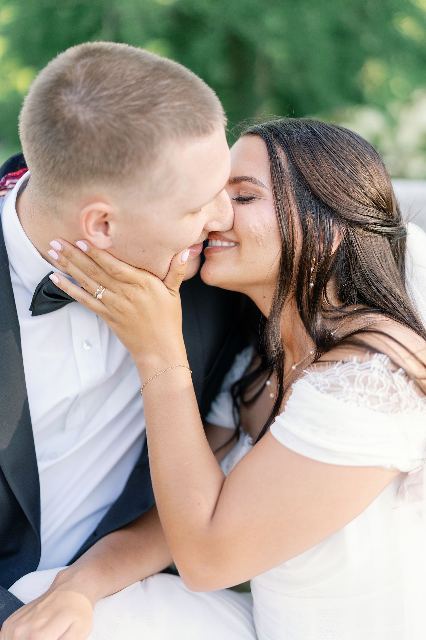 A bride and groom share a romantic kiss during their wedding day, with the bride in a white off-shoulder gown and a diamond ring.