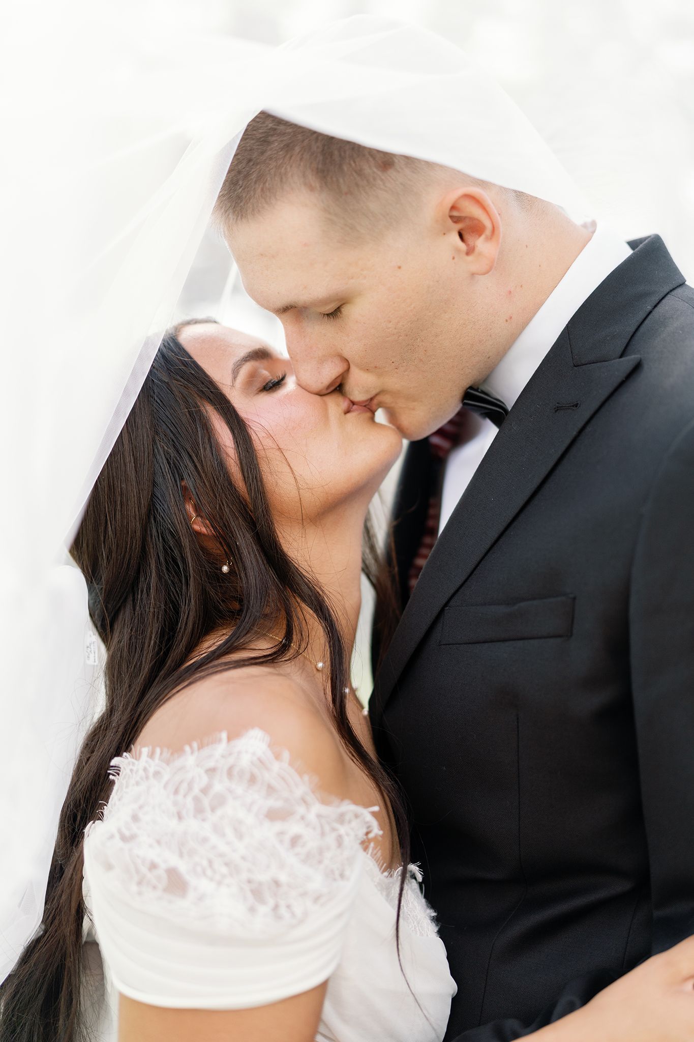 Bride and groom sharing a romantic kiss under a flowing white veil on their wedding day.