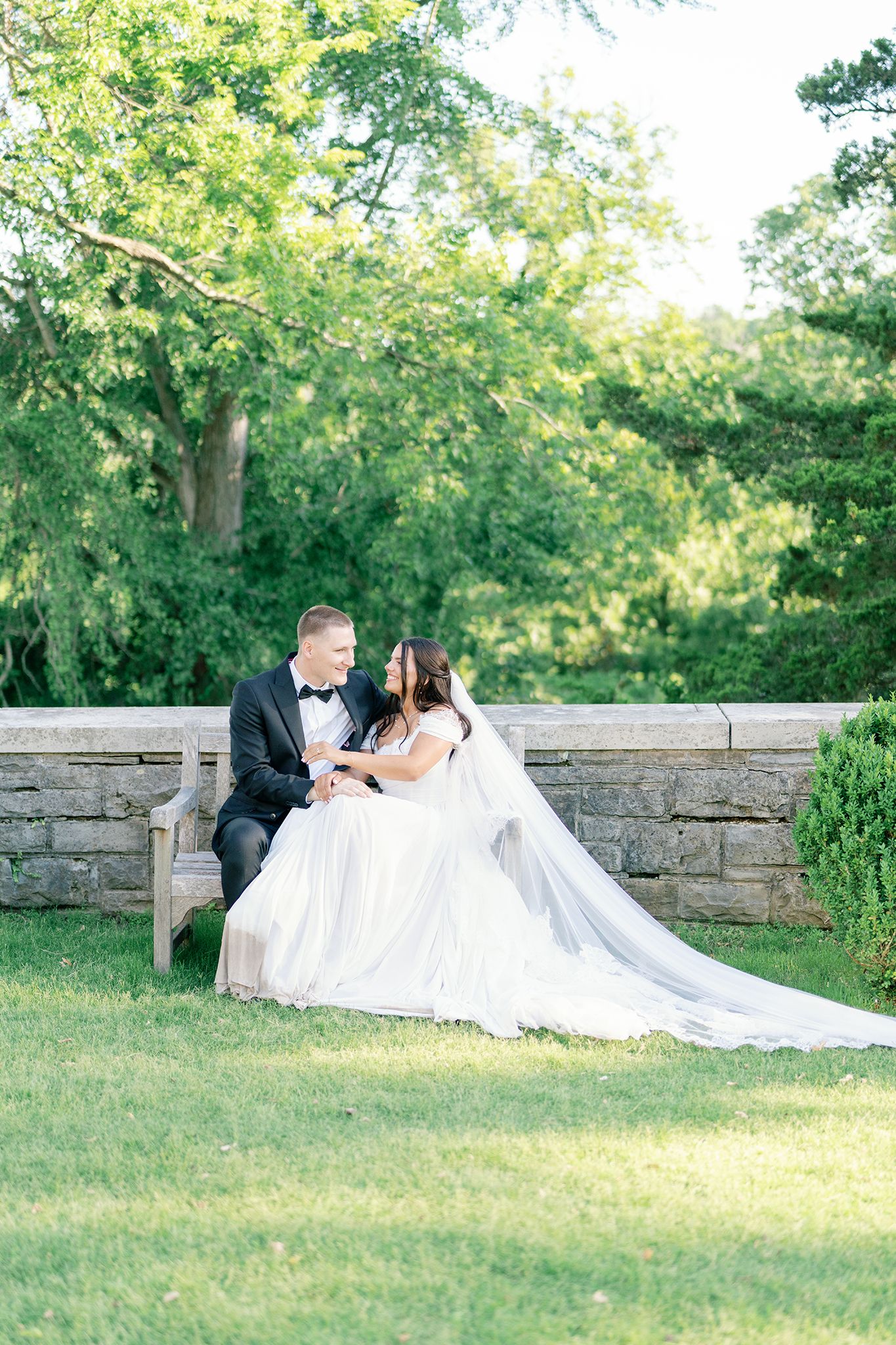 Bride and groom sit together on a wooden bench in a lush garden, sharing an intimate moment in wedding attire.