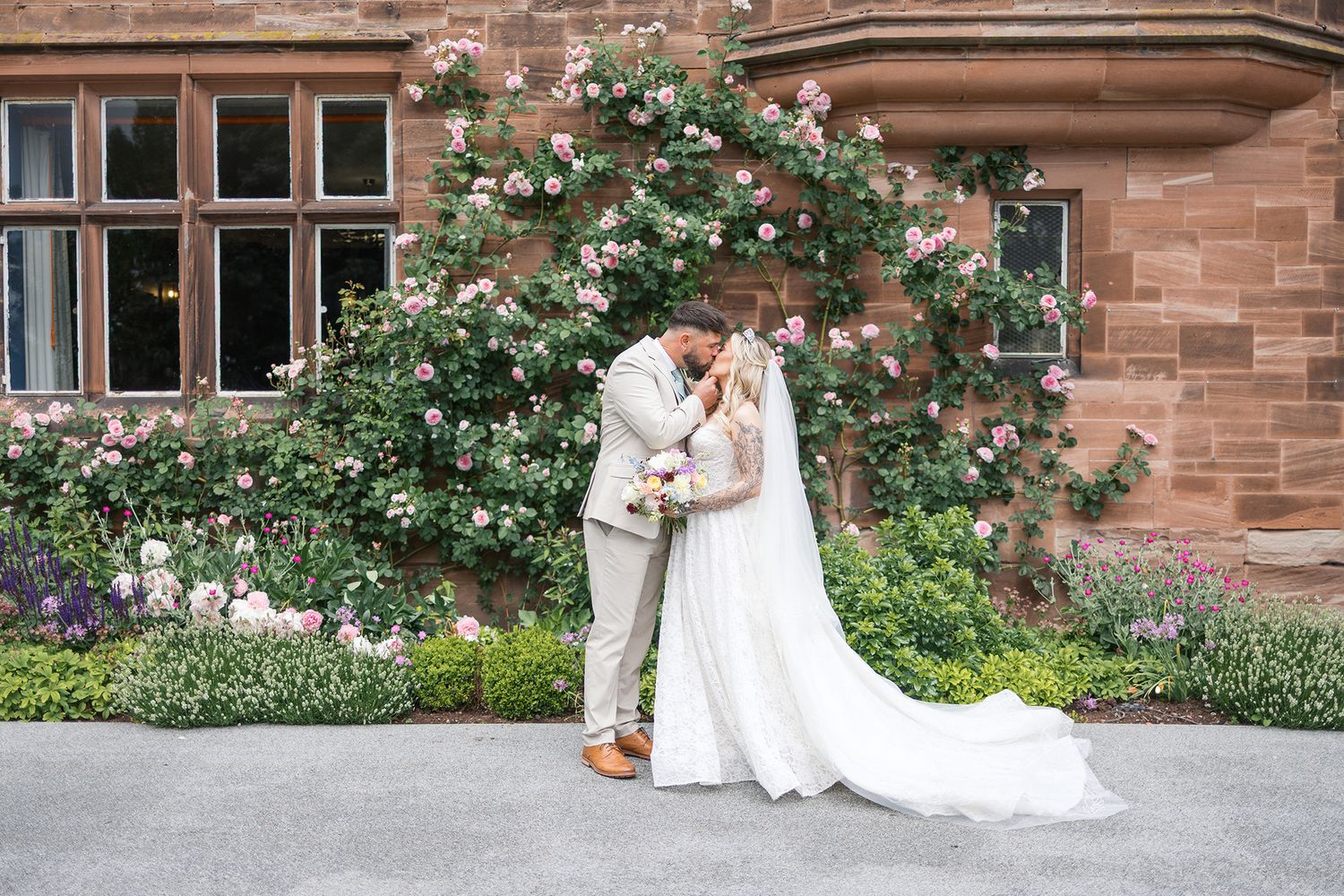 Garden photoshoot at historic brick building featuring white dress against climbing roses and stone architecture.