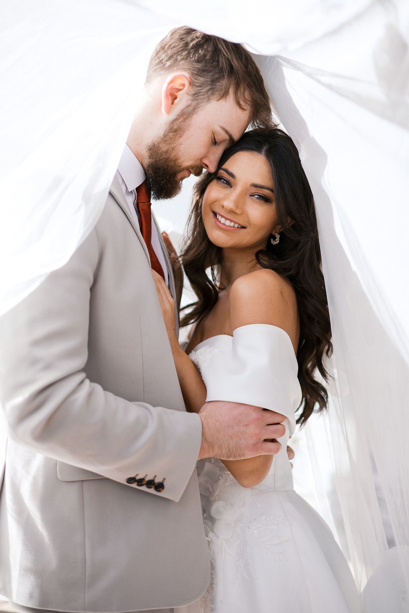 Joyful bride in white dress and groom in beige suit embrace beneath sheer white veil during wedding photoshoot.