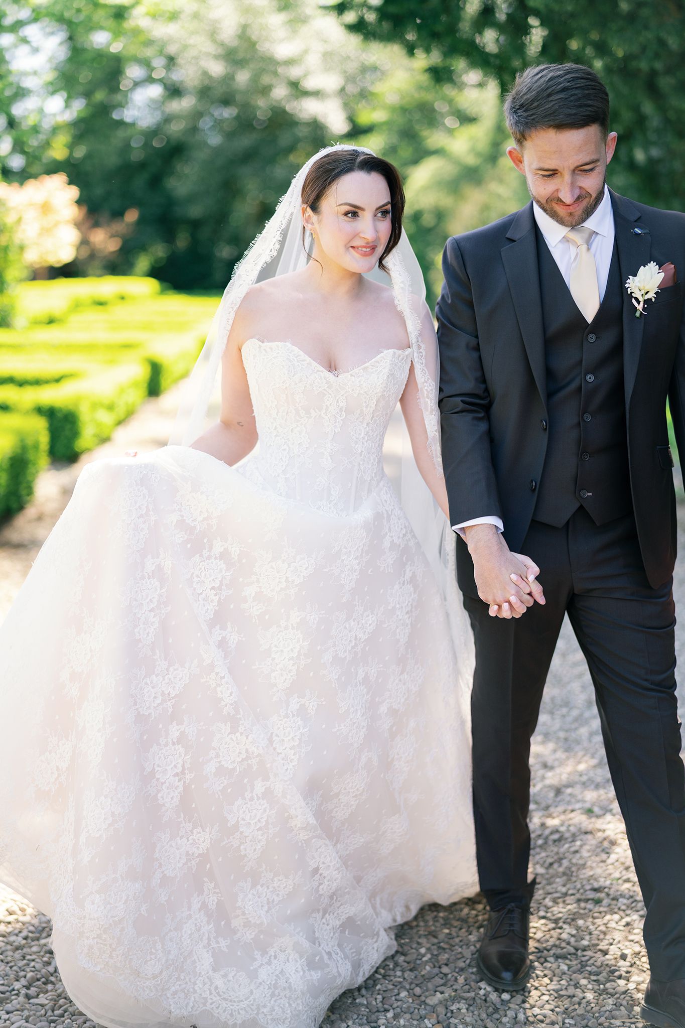 Bride and groom walking hand in hand in a garden, bride wearing a lace ballgown and veil.