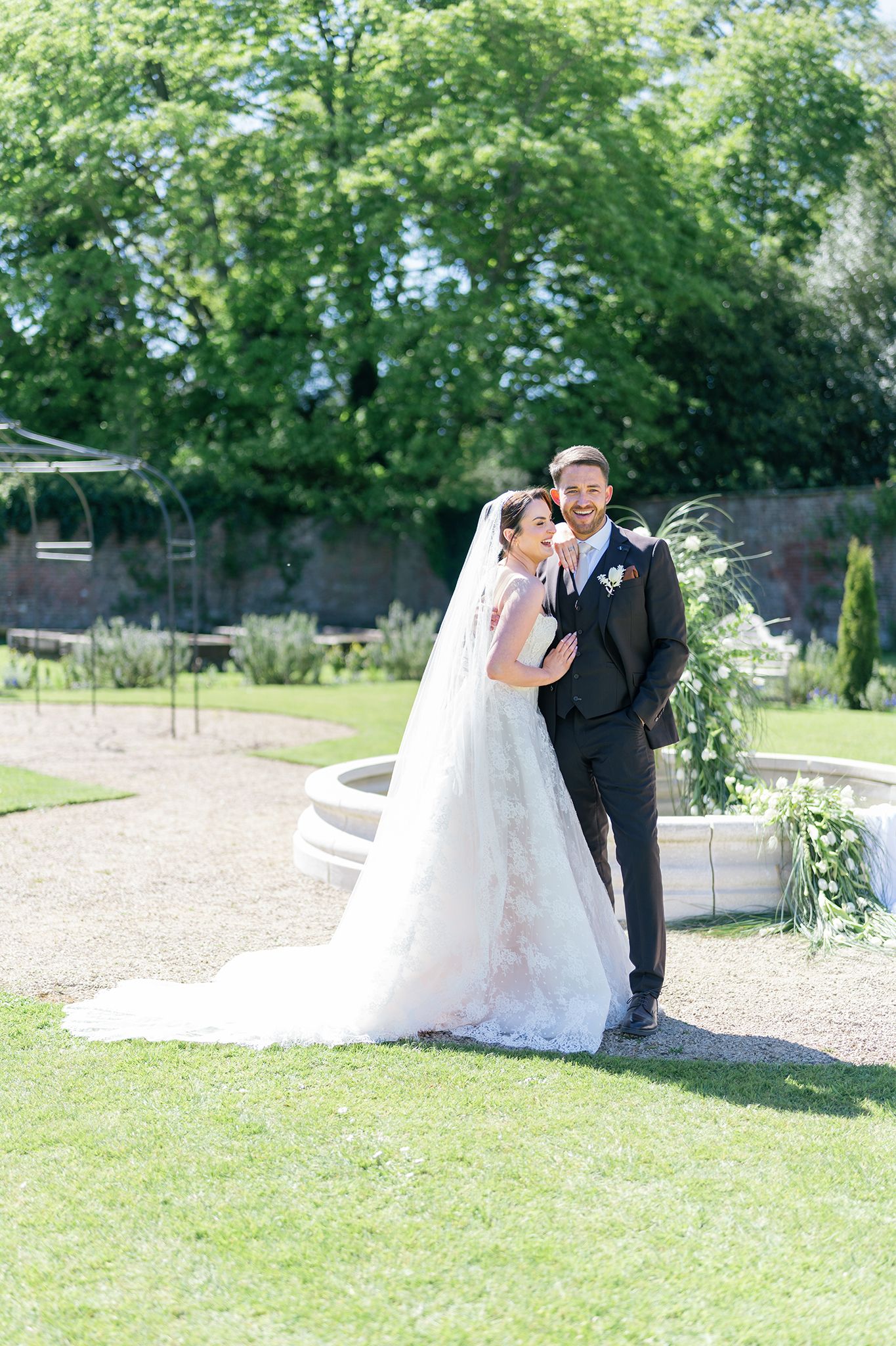 Bride in white gown and veil stands with groom in black suit in a sunlit garden with fountain backdrop.