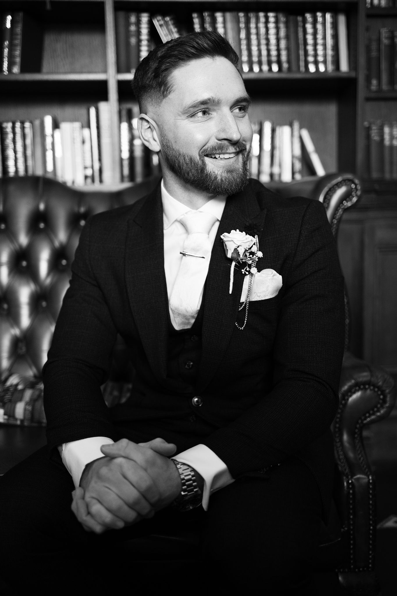 Black and white portrait of a bearded groom in a three-piece tuxedo sitting in a leather chair with bookshelves behind him.