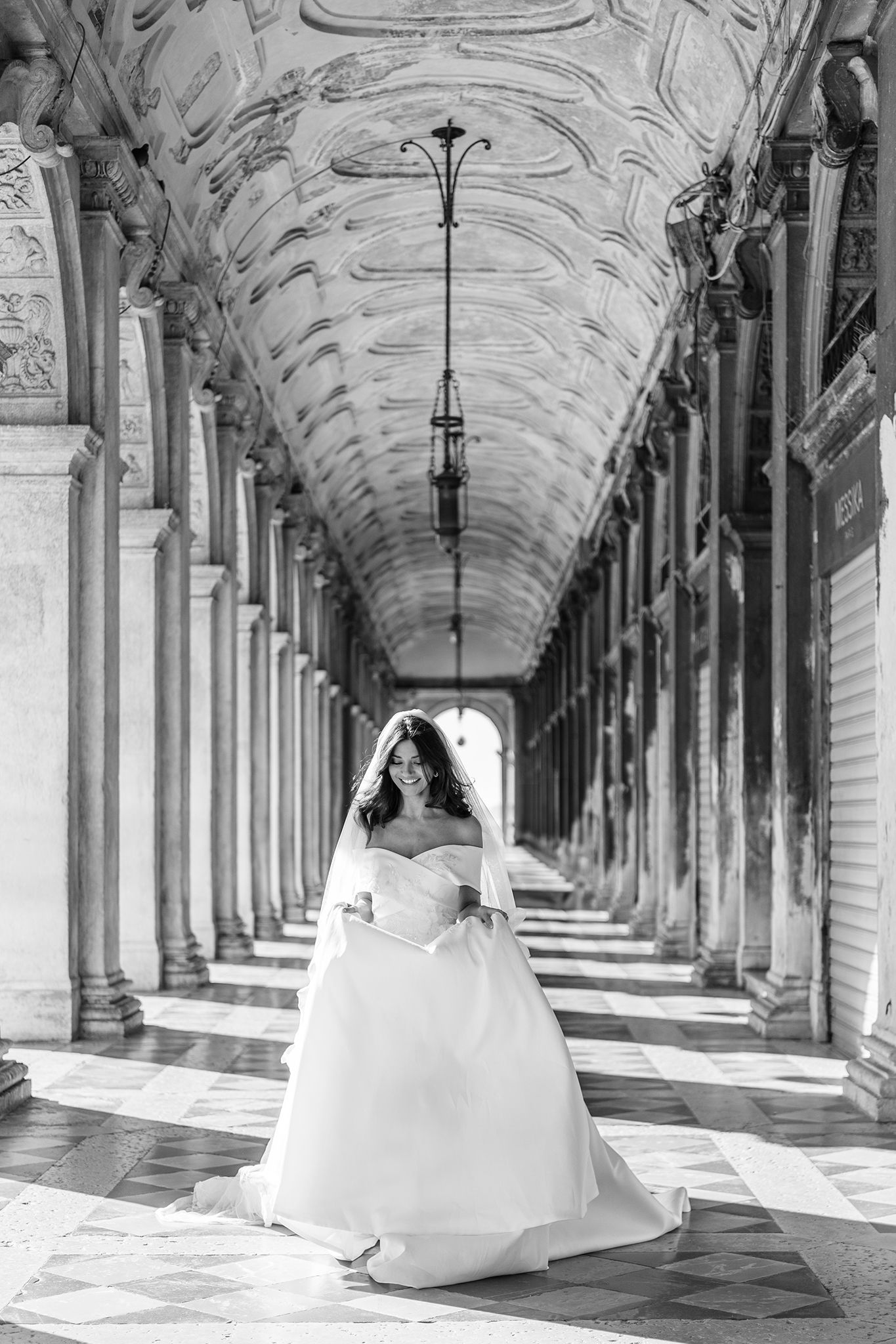 A bride in a white wedding gown stands in an ornate covered colonnade with classical architecture and natural lighting.