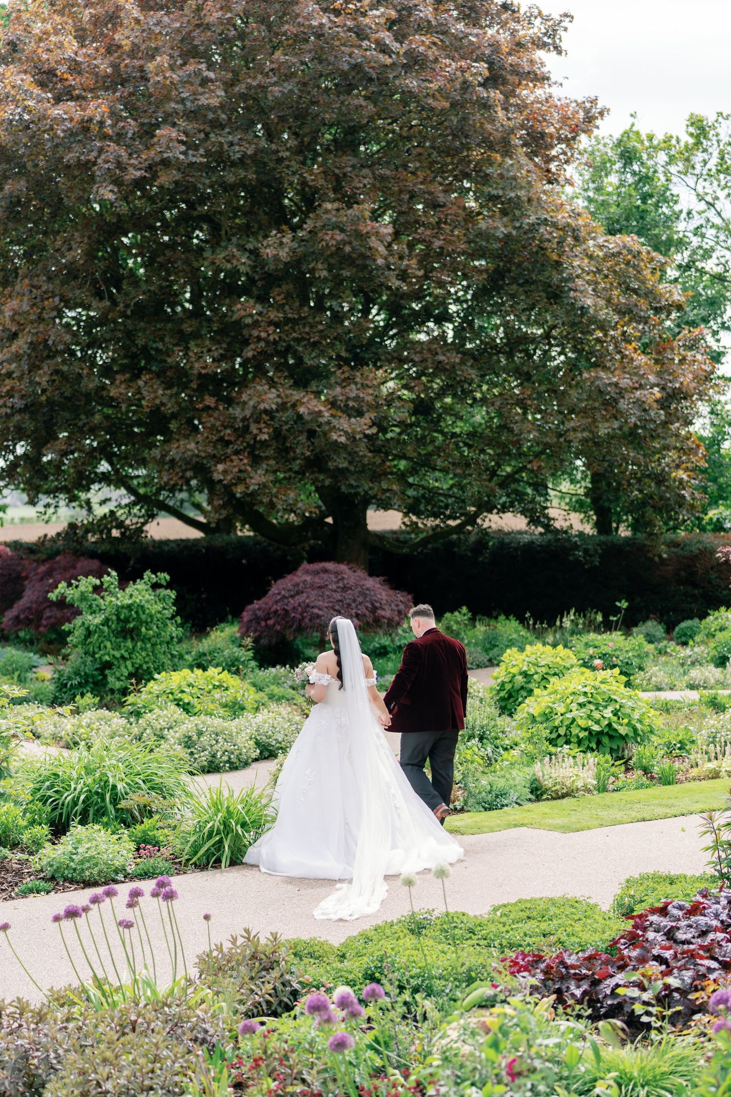 Bride and groom walking hand in hand through a lush garden with colorful flowers and a large tree.