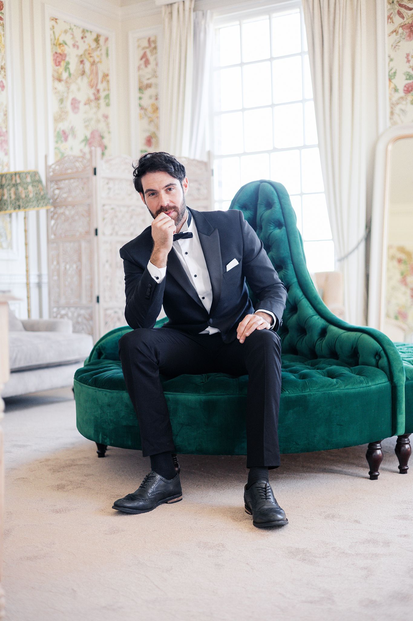 Elegant tuxedo-clad figure poses thoughtfully on emerald green velvet chaise lounge in luxurious room with floral wallpaper.
