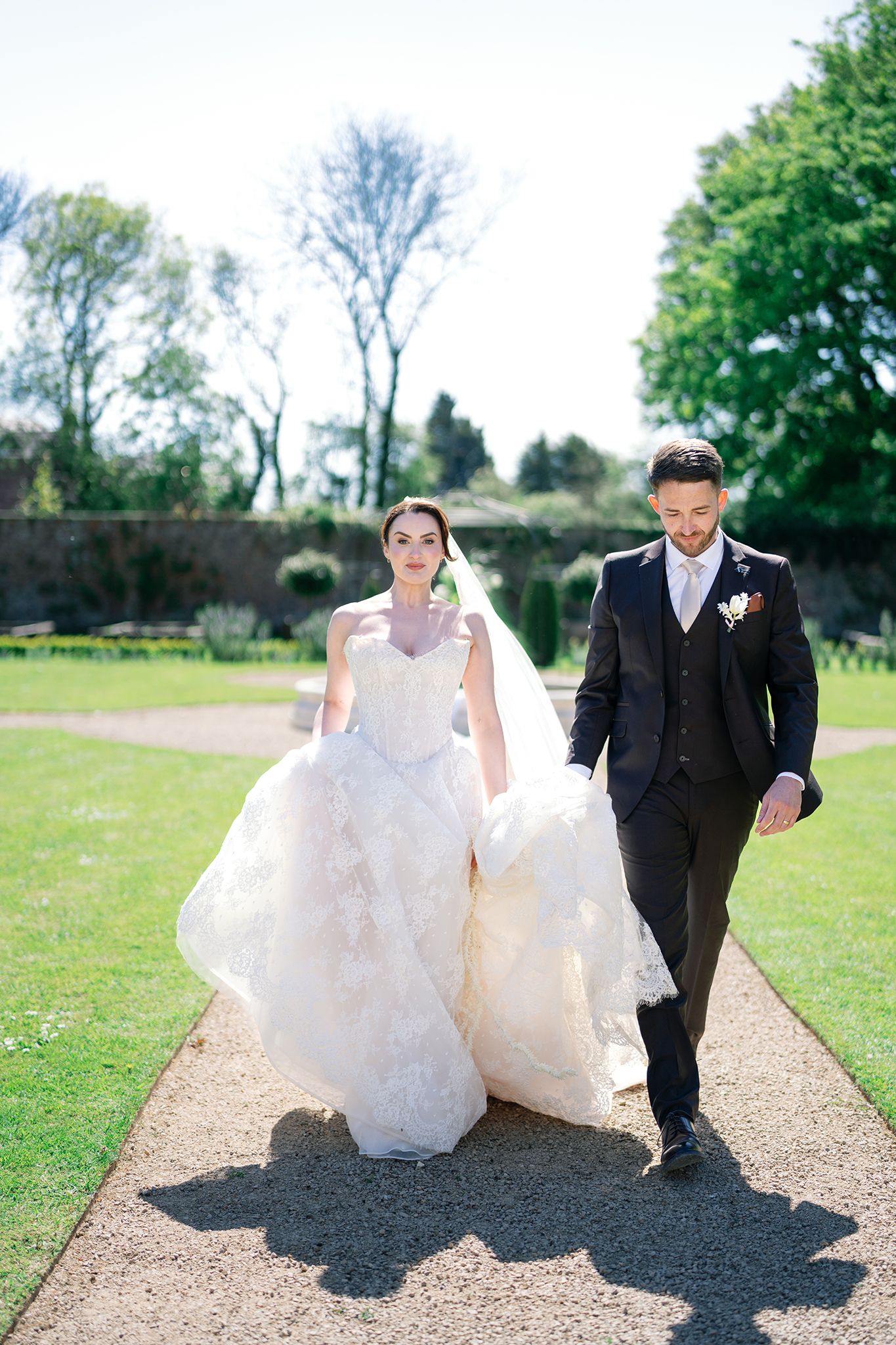 Bride in white ballgown and groom in dark suit walk hand-in-hand through a sunlit garden path on their wedding day.