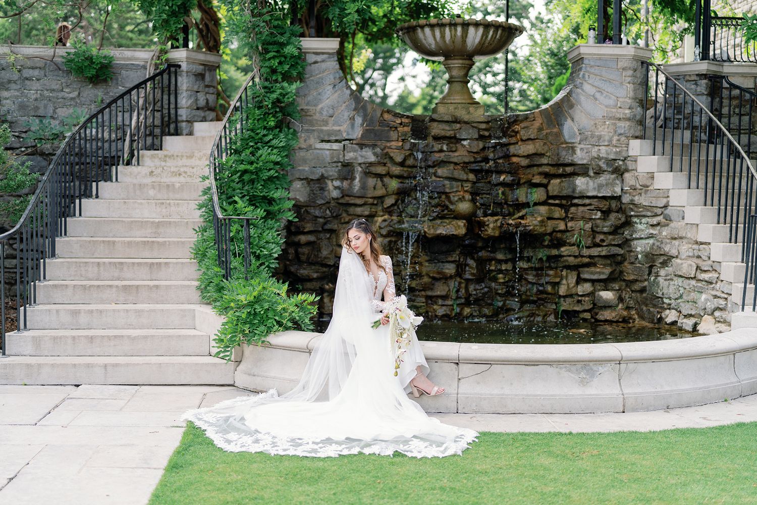 Bride in white gown and veil poses by stone fountain with dual staircases in lush garden setting.