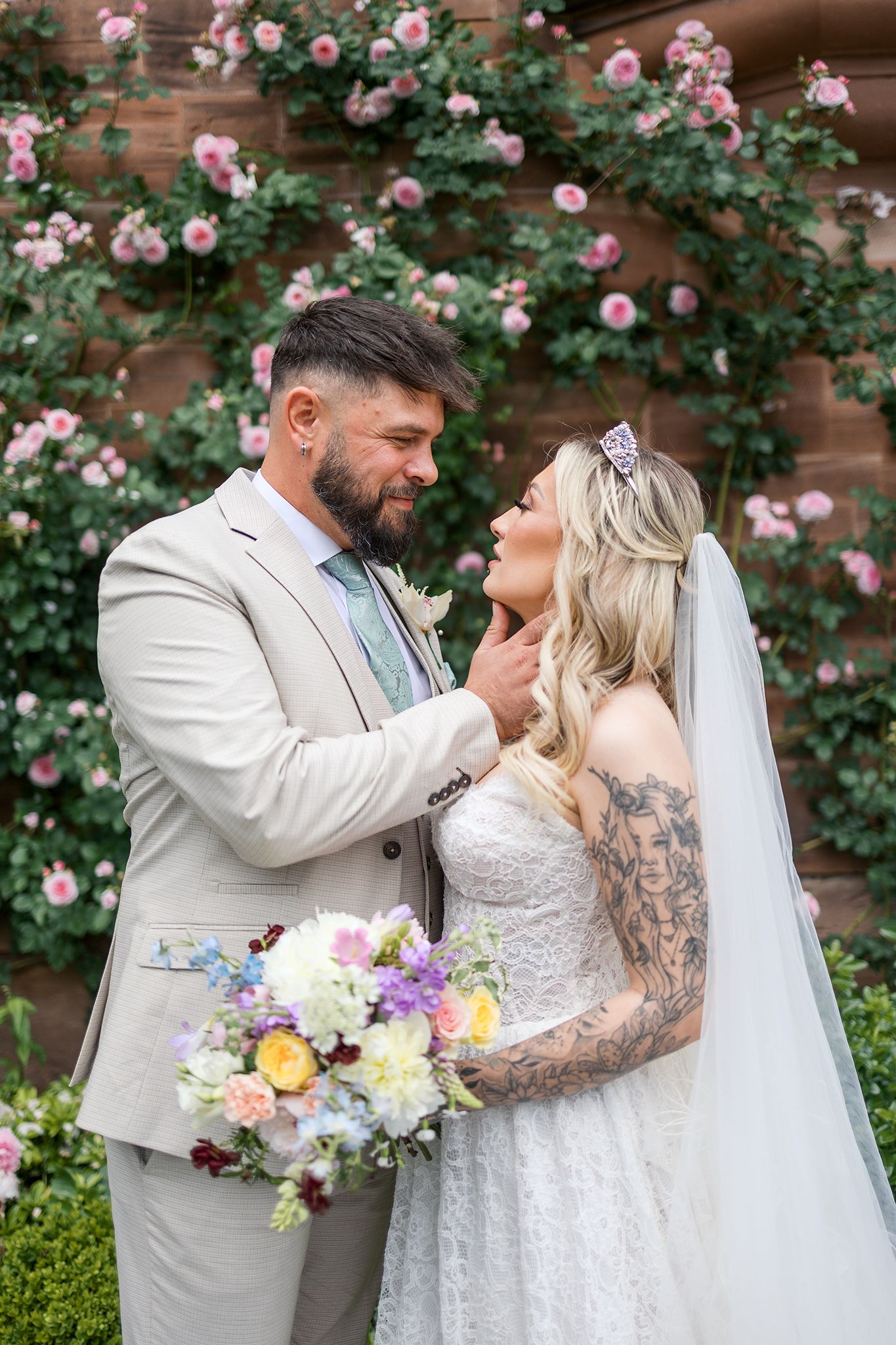 Romantic garden wedding moment with pink roses climbing on wooden wall behind the couple in light formal attire.