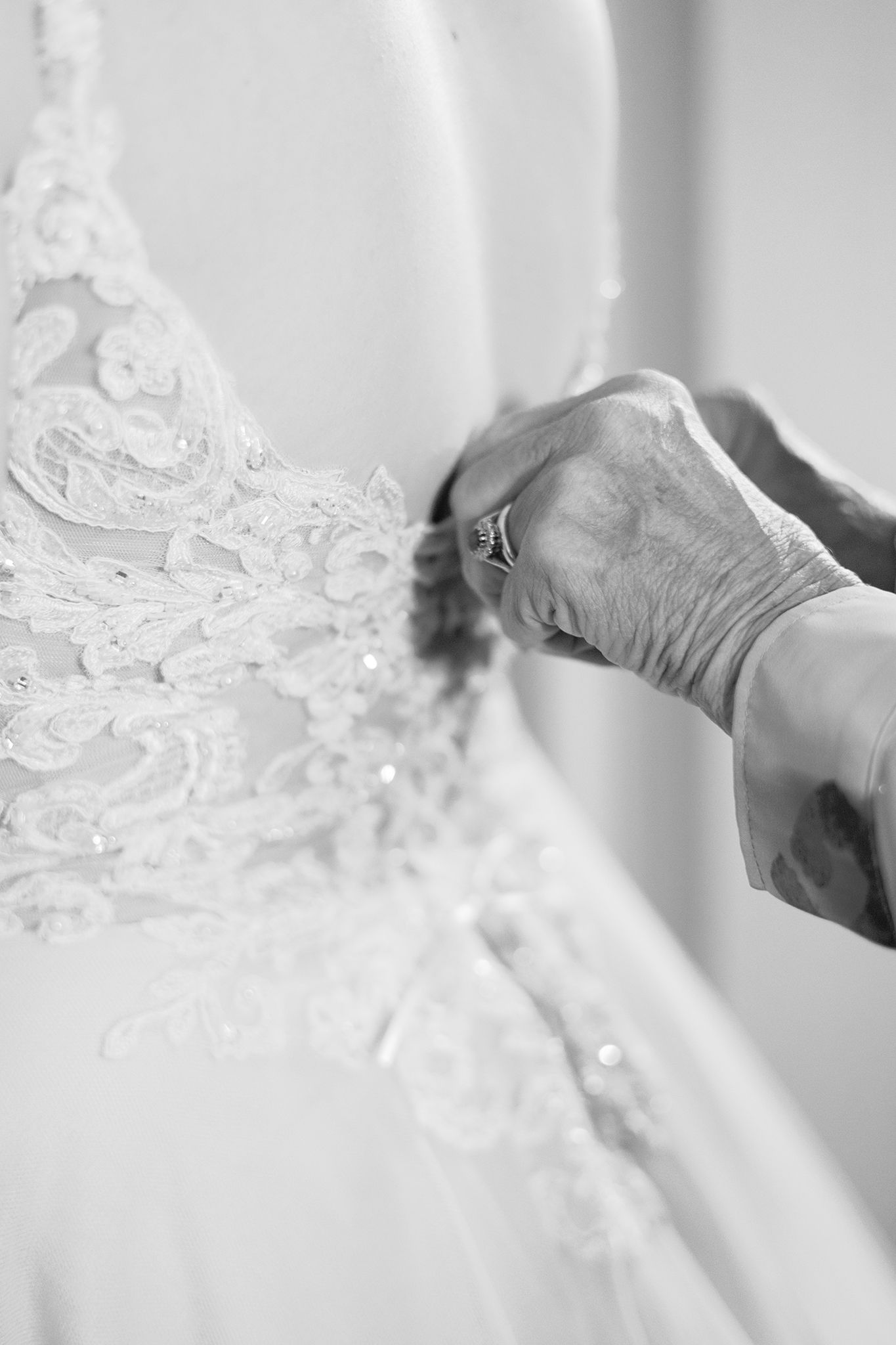 Close-up black and white detail of delicate lace and beading on a wedding dress being adjusted by hands.