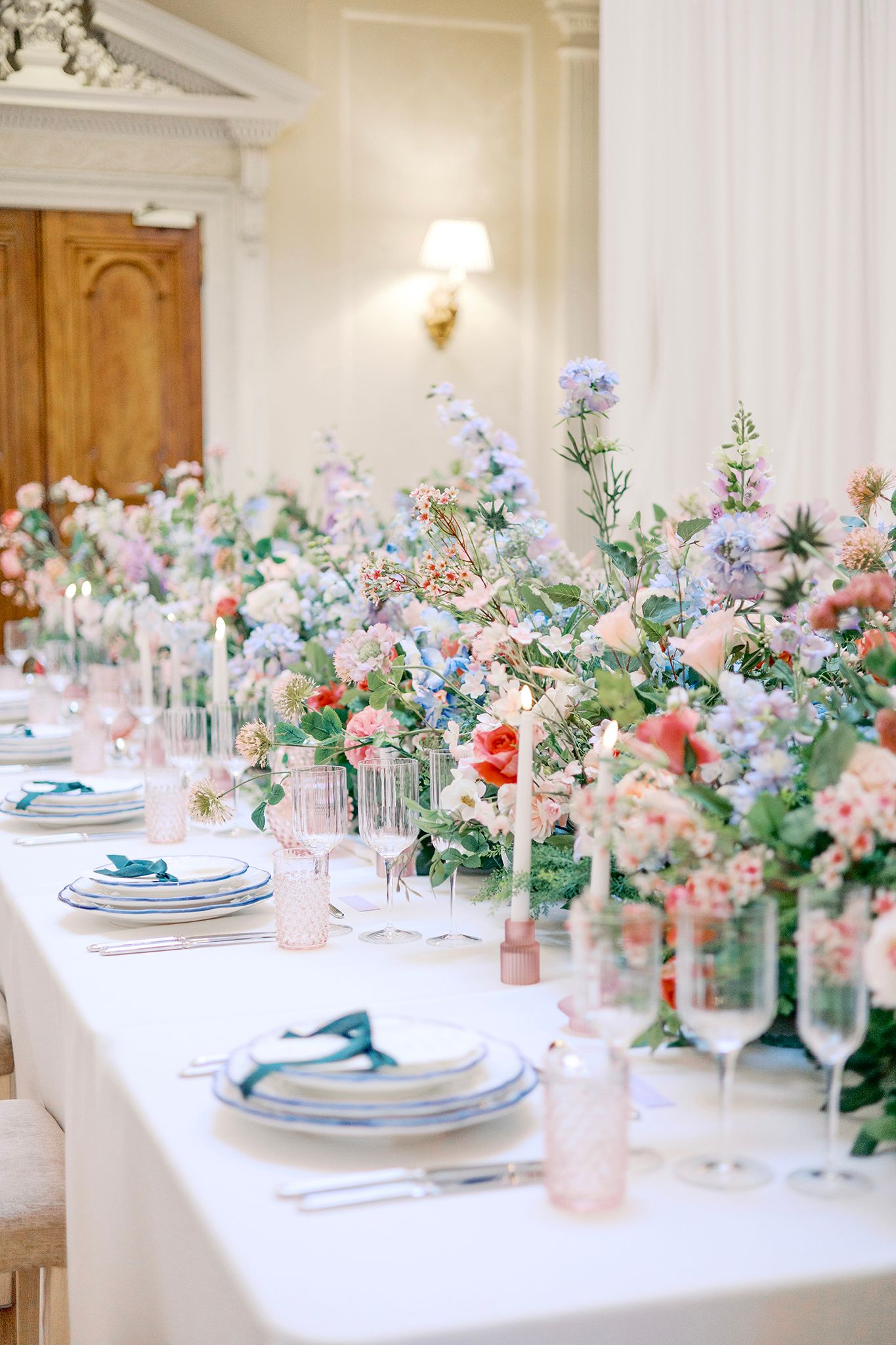 Elegant wedding table setting with pastel floral centerpieces featuring blush and blue flowers in glass vases along white tablecloth.