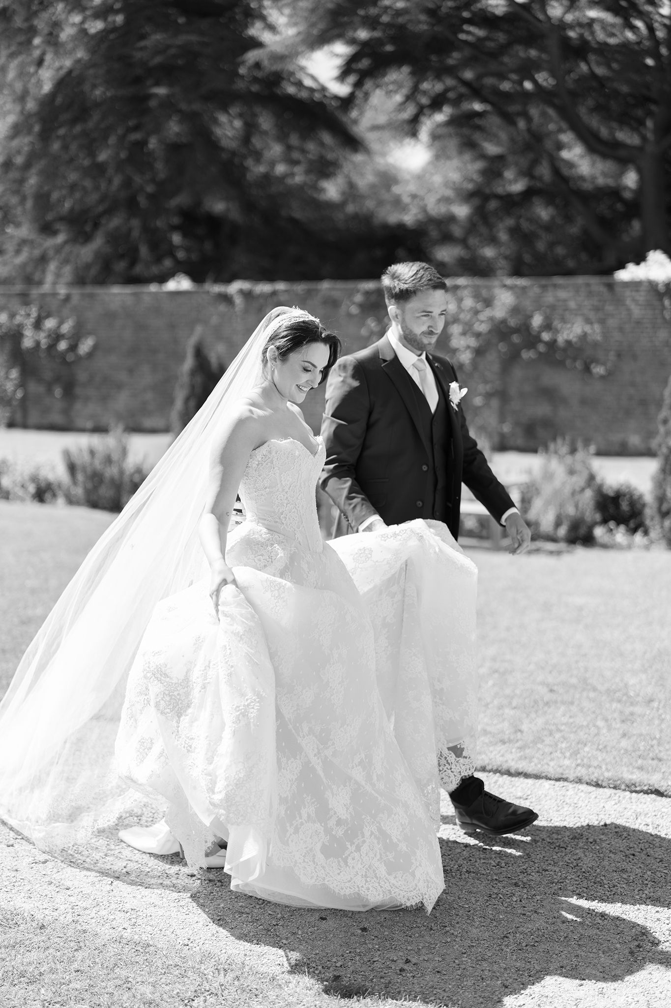 Black and white photo of a bride and groom walking outdoors, the groom helping carry her lace wedding dress train.