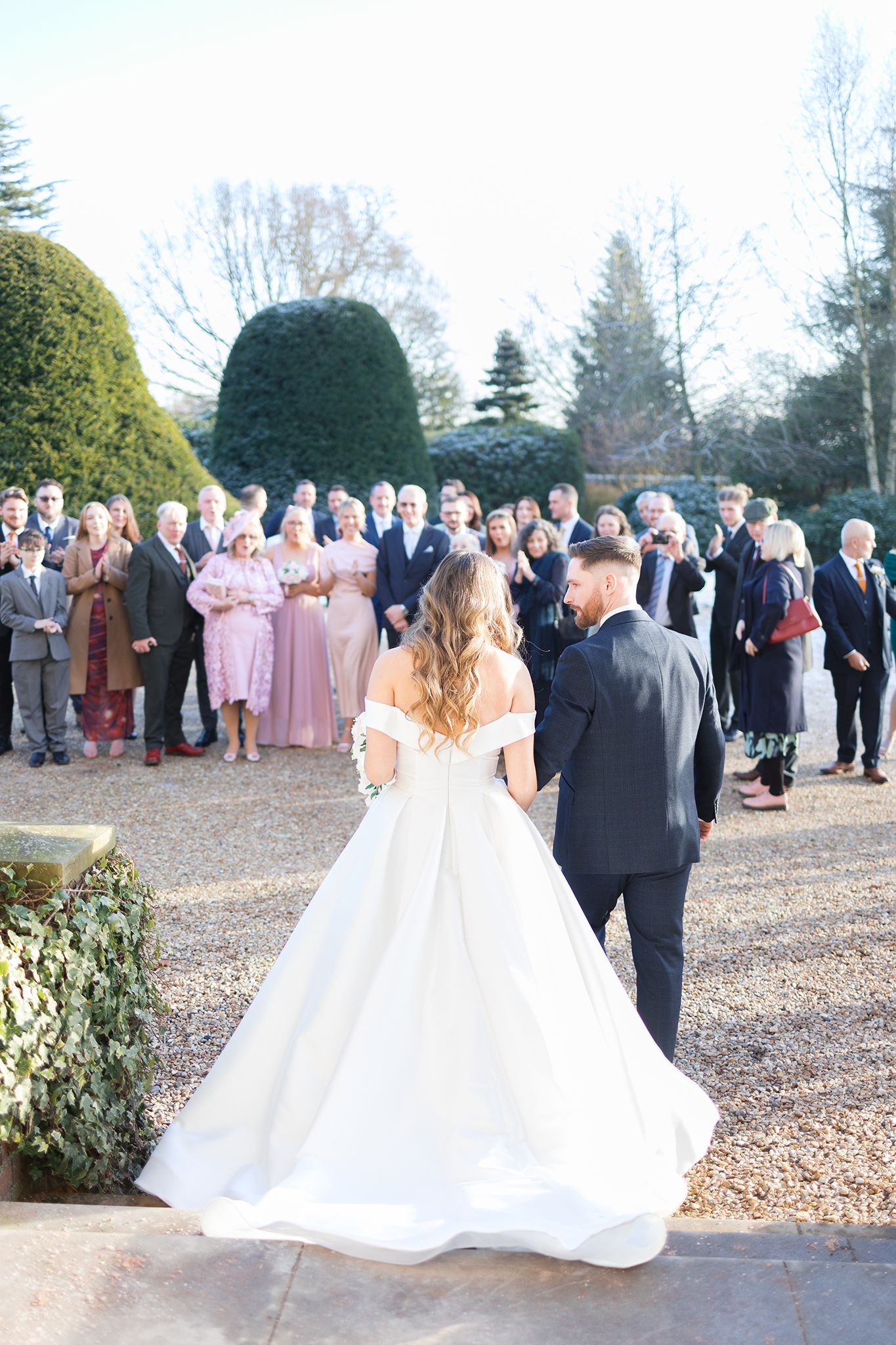 Wedding guests gather to watch as couple stand together near manicured garden hedges on their special day.