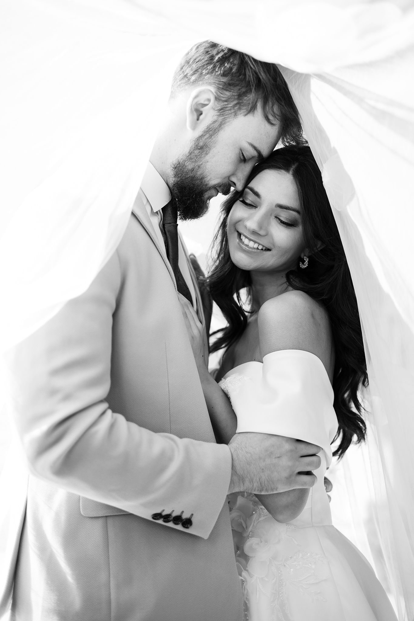 A tender black and white wedding portrait showing a couple embracing under a veil, sharing an intimate moment together.
