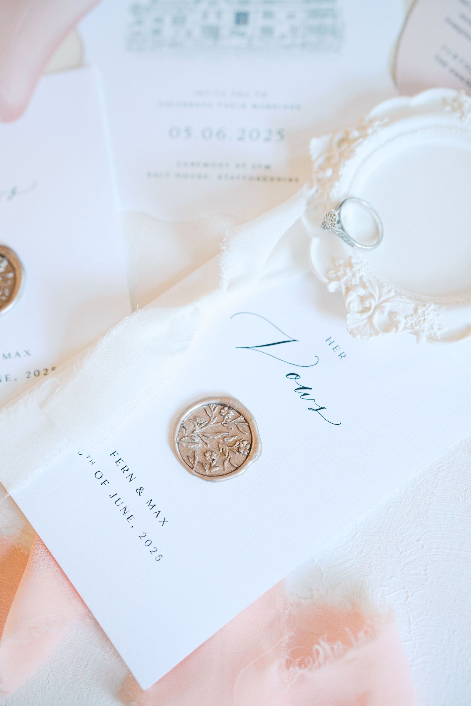 Elegant wedding stationery flat lay with wax seal vow booklet, diamond ring, and soft pink feathers on white background.