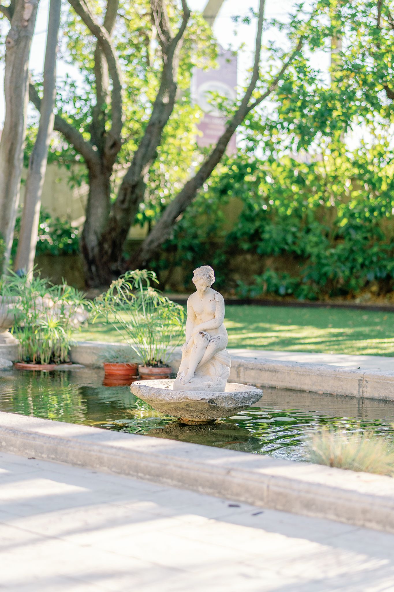 A classical stone statue of a figure sits atop a fountain in a lush green garden with trees and potted plants.
