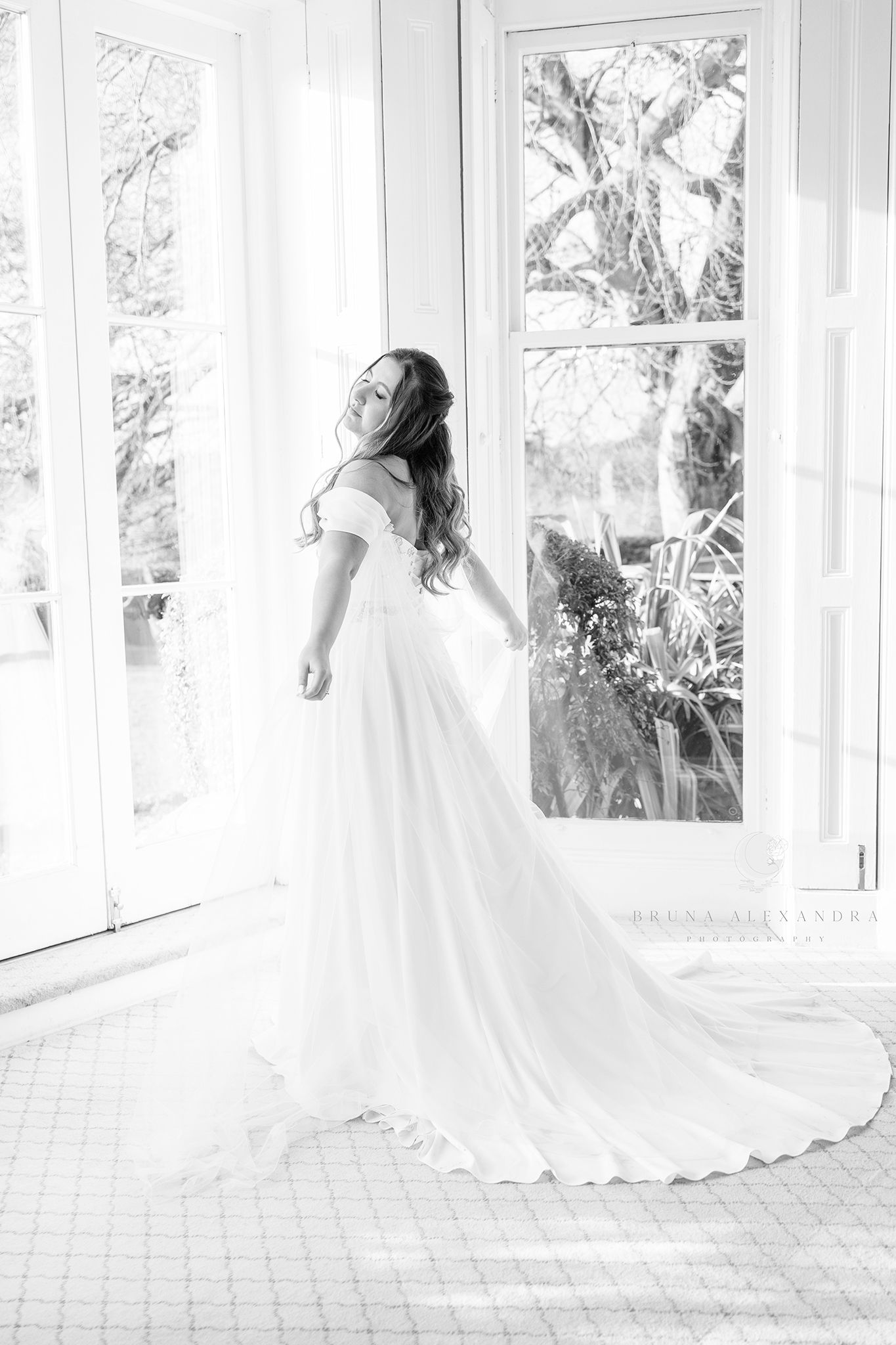 Ethereal black and white bridal portrait near sunlit windows showing flowing wedding gown.