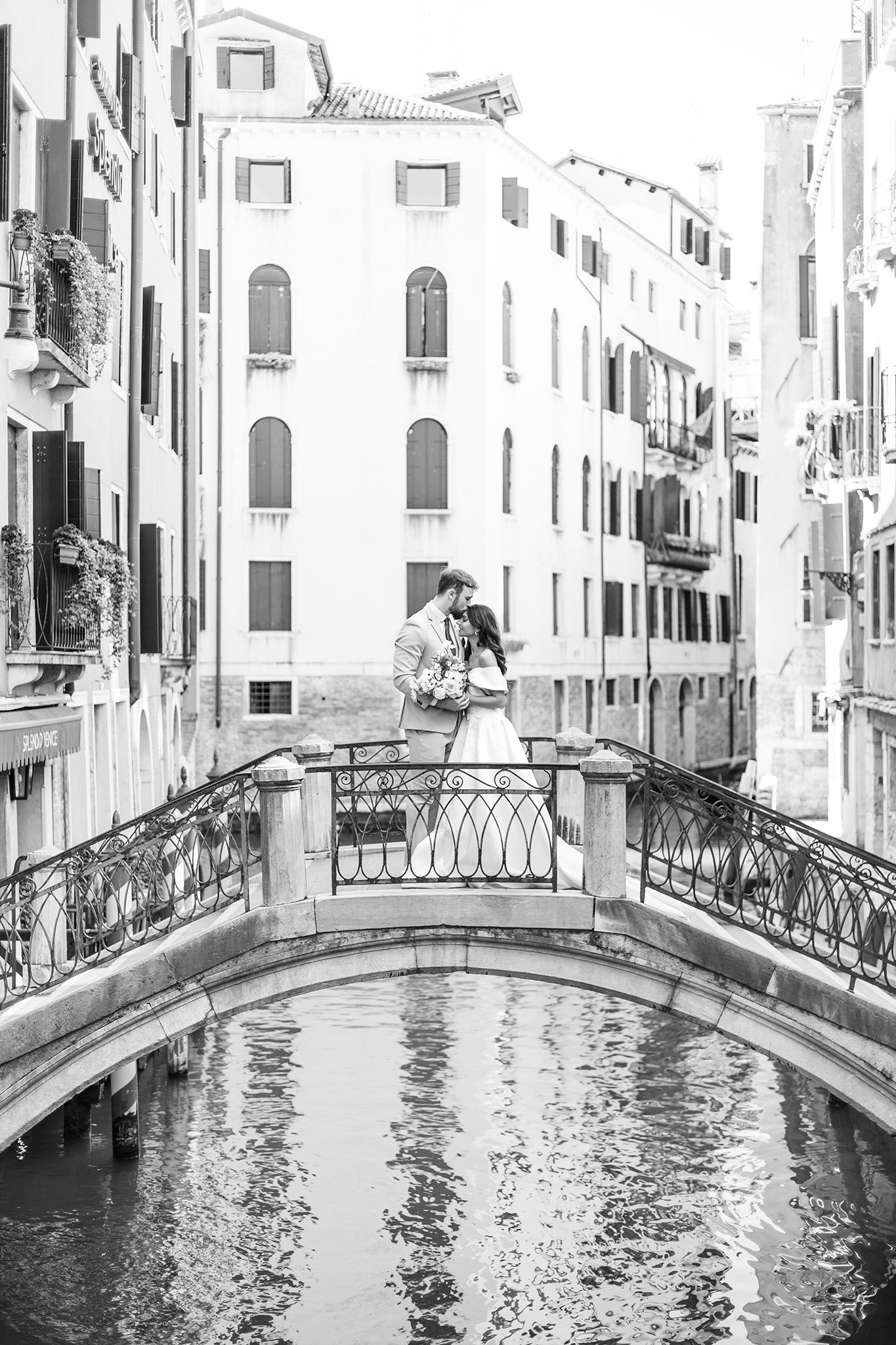Black and white photo of couple sharing an umbrella on an ornate bridge over a scenic Venice canal with historic buildings.