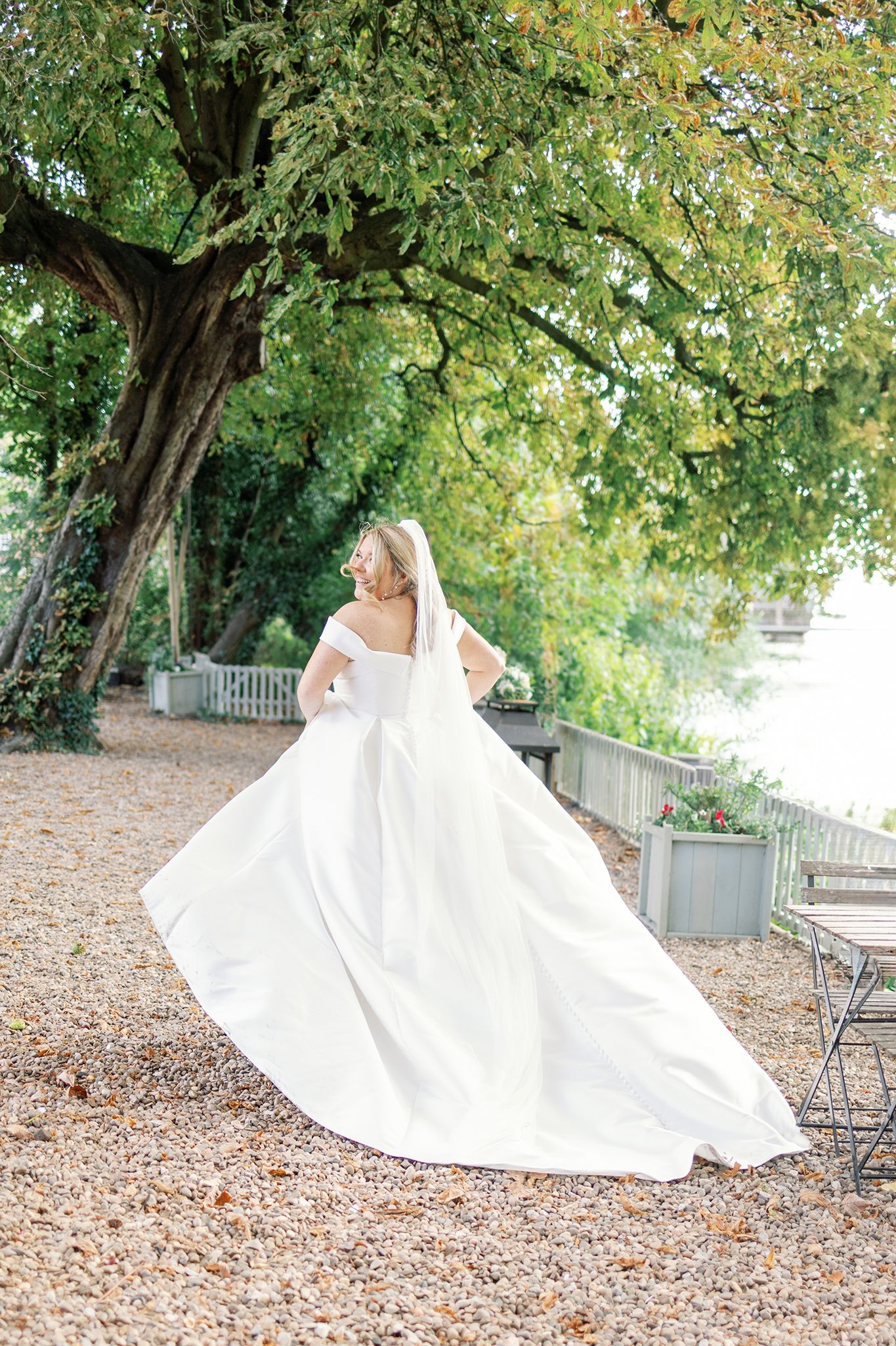 A flowing white wedding dress twirls gracefully on fallen autumn leaves under trees in an outdoor garden setting.