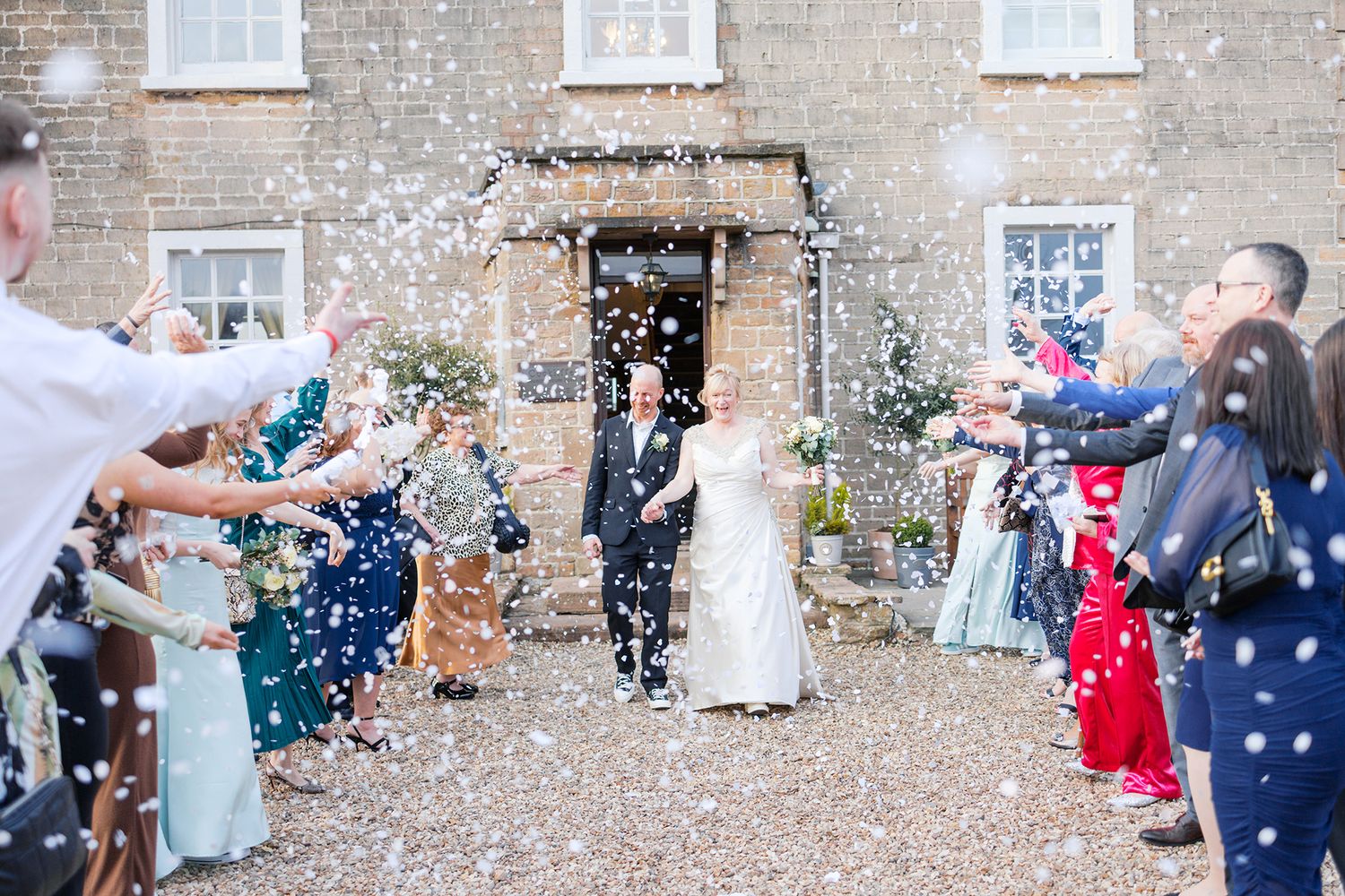 Wedding guests throw confetti over couple exiting stone building as they celebrate at countryside venue.