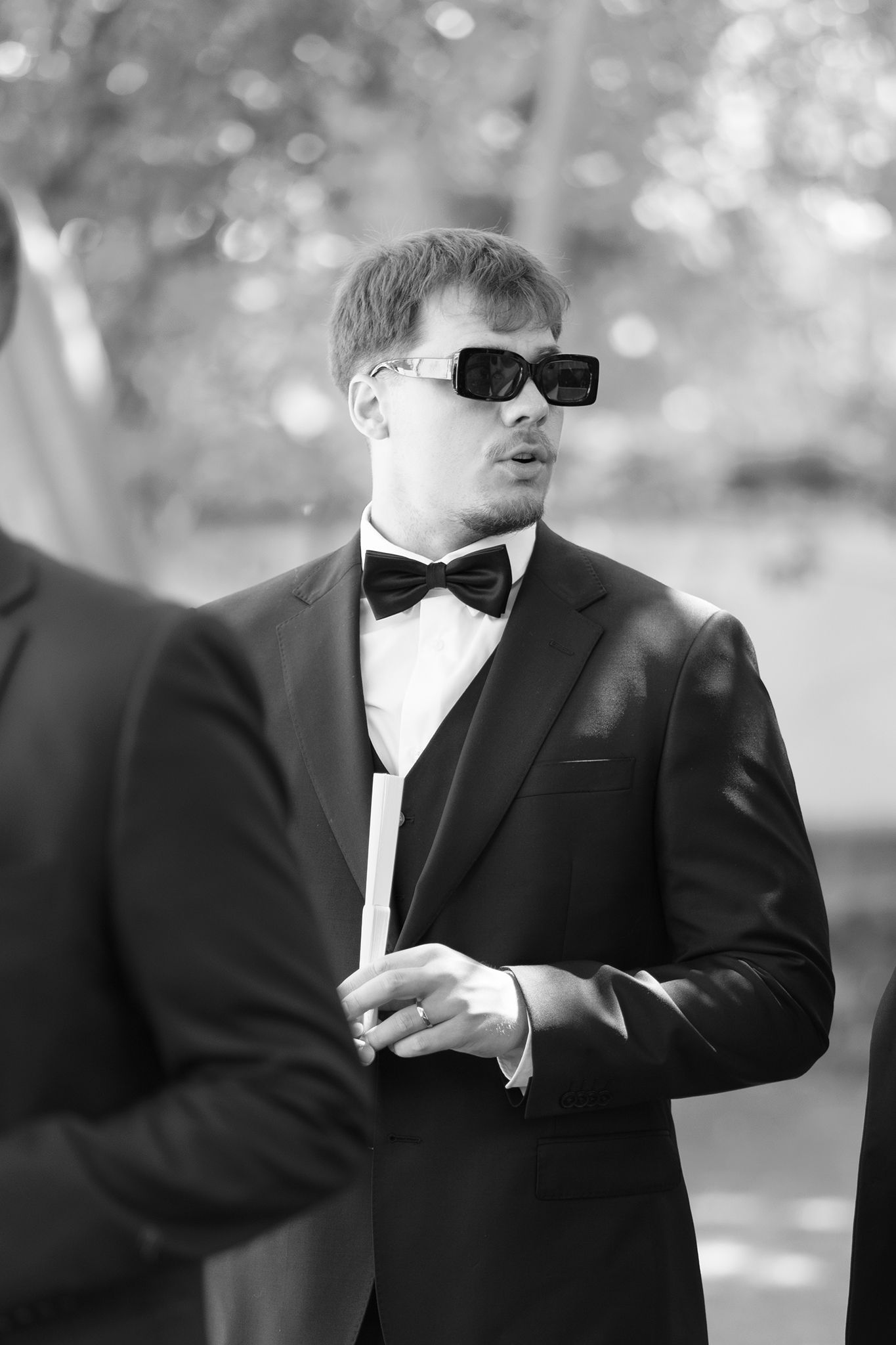 Black and white photo of a stylish man in a tuxedo and bold sunglasses at an outdoor formal event.