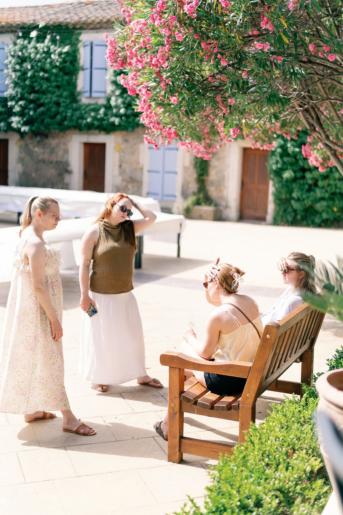 Four women socializing outdoors near a flowering tree beside a rustic stone building on a sunny day.