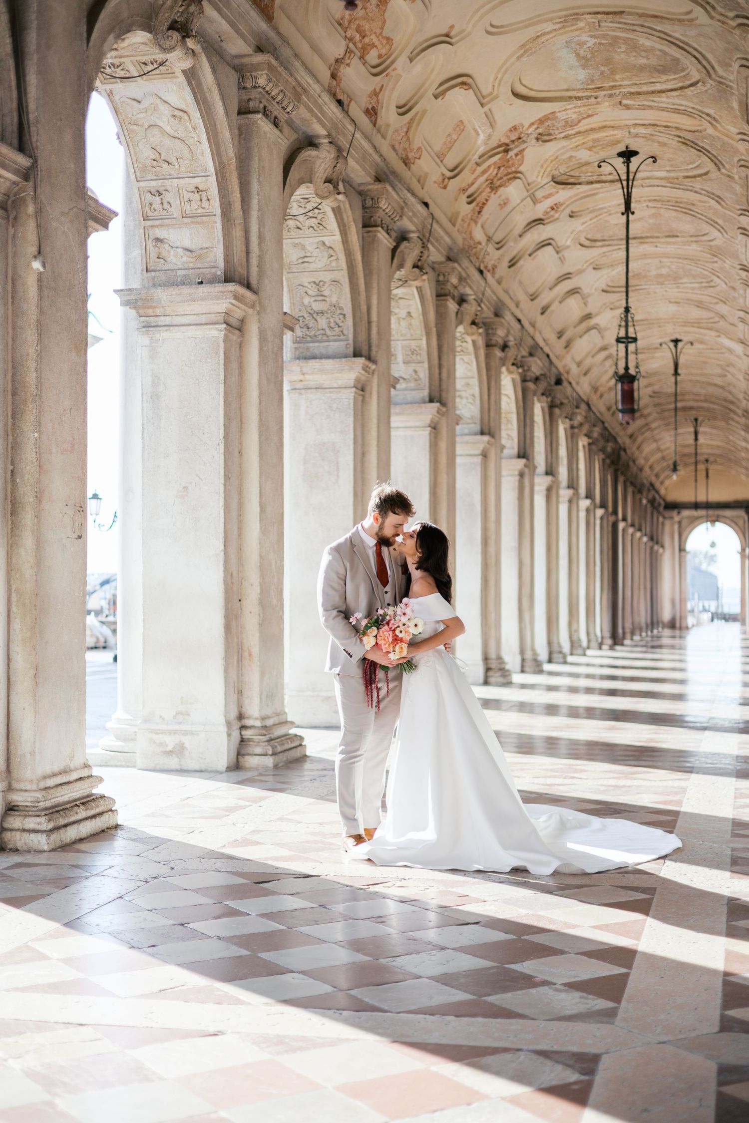 Bride and groom share a romantic kiss under ornate marble arches in Venice, Italy, bride holding a pink bouquet.