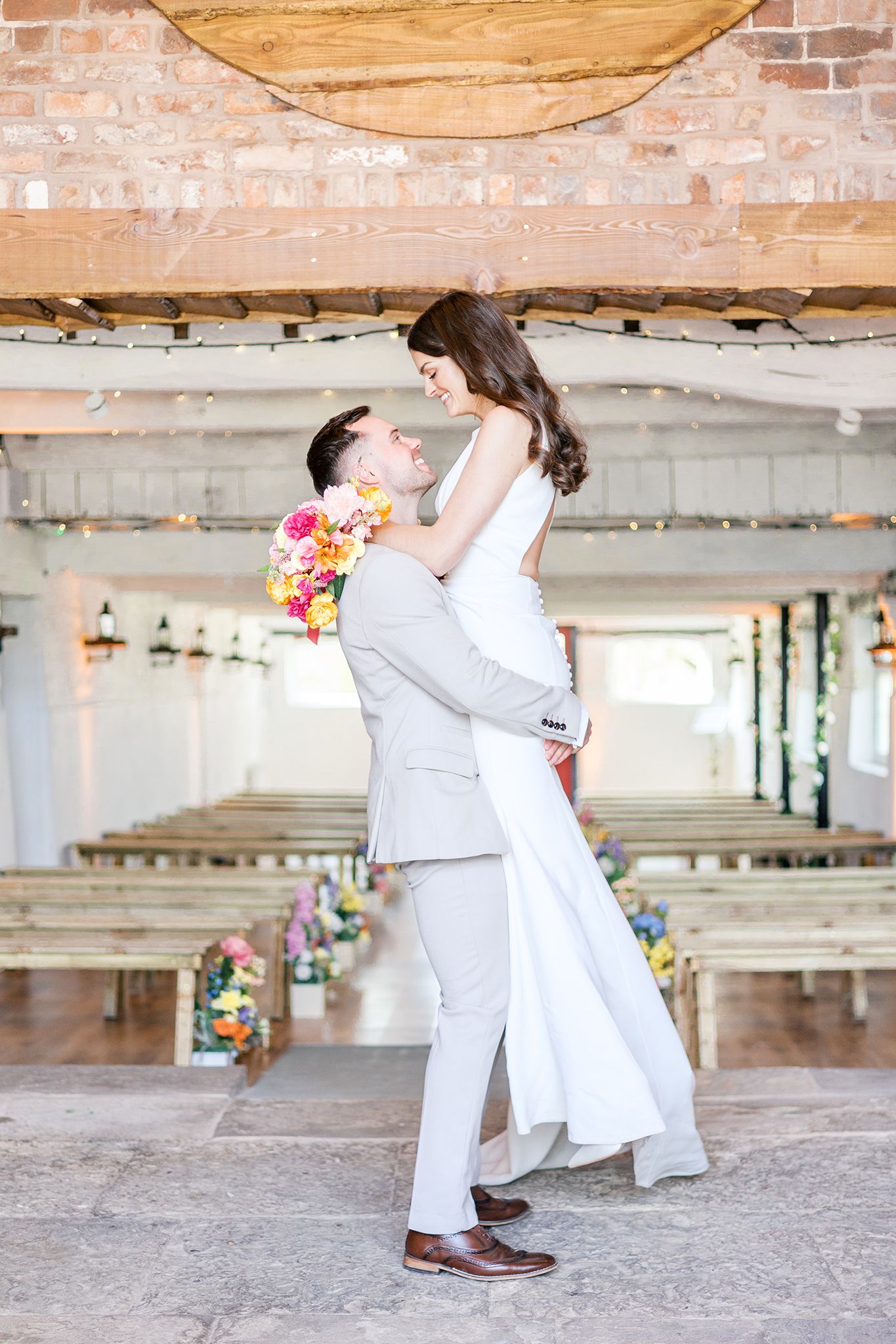 A romantic wedding couple embraces in an industrial venue with exposed brick walls and colorful floral bouquet.