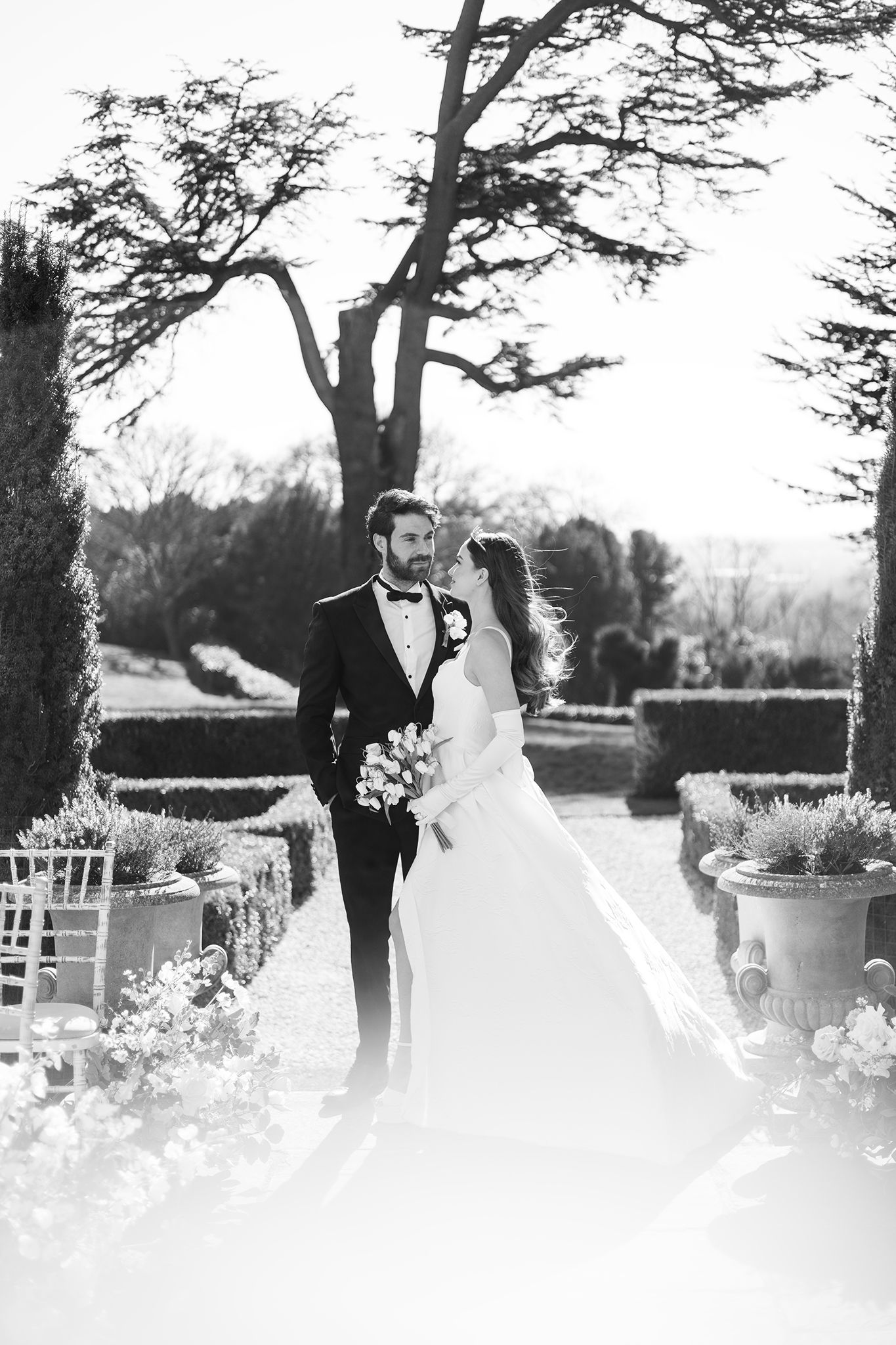 A romantic black and white wedding photo in a garden setting with elegant trees and white chairs along the aisle.