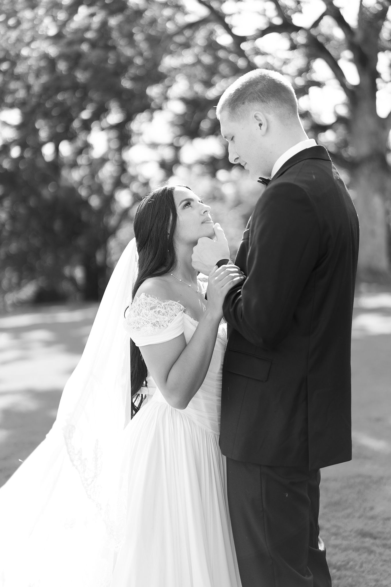 Black and white photo of a bride and groom holding hands and gazing at each other outdoors on their wedding day.