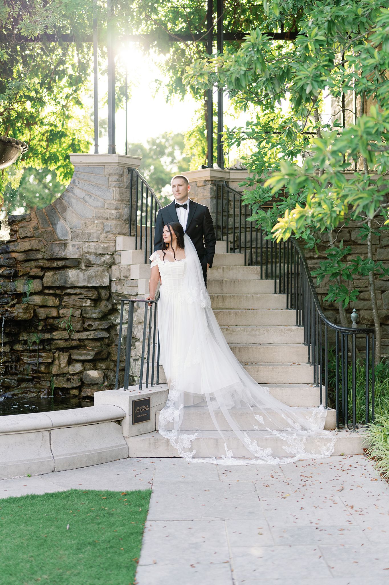 Bride and groom pose on stone steps surrounded by lush greenery, bride wearing a flowing lace cathedral veil.