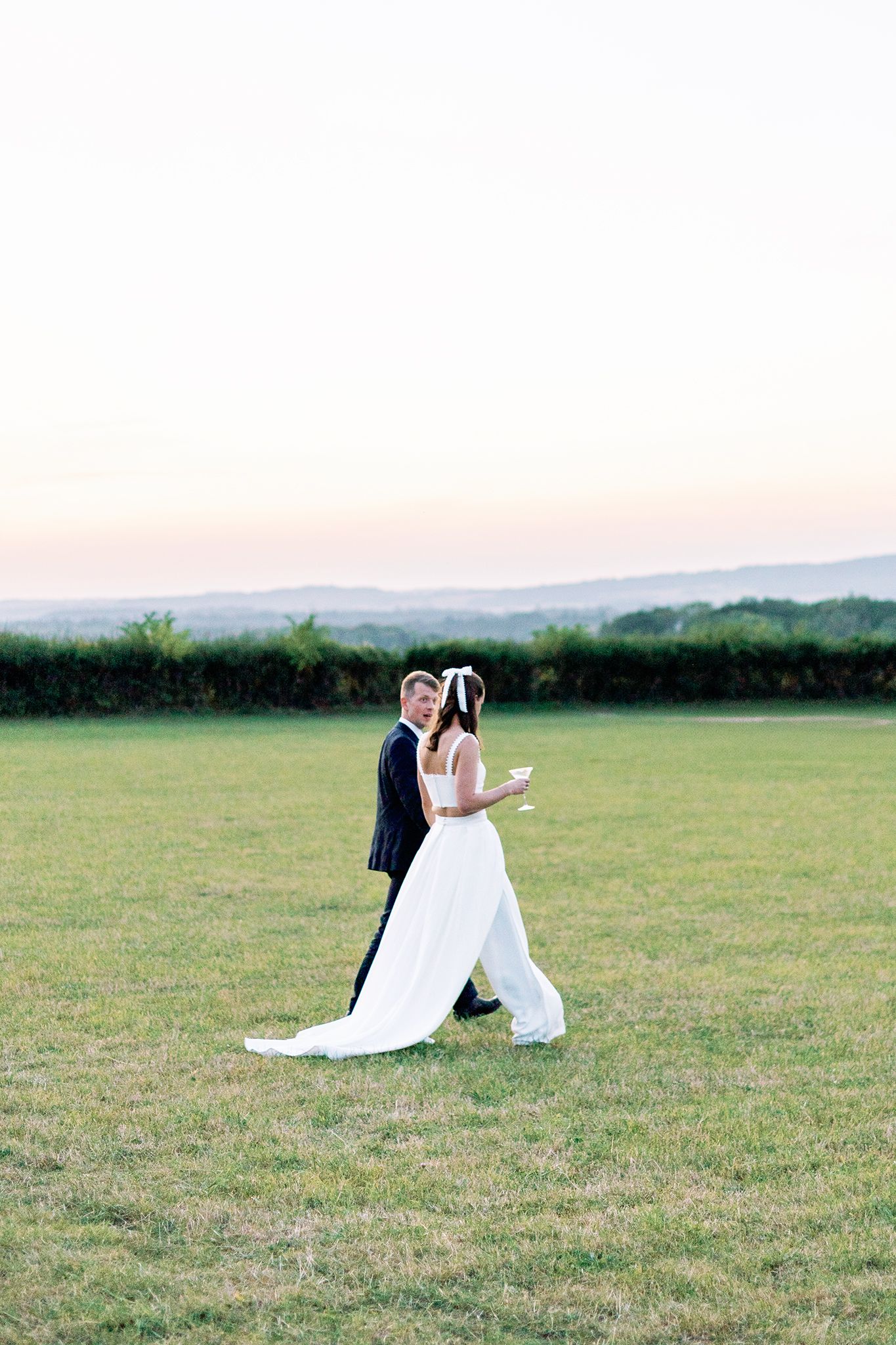 A couple in formal wedding attire share a romantic moment in a scenic field at sunset.