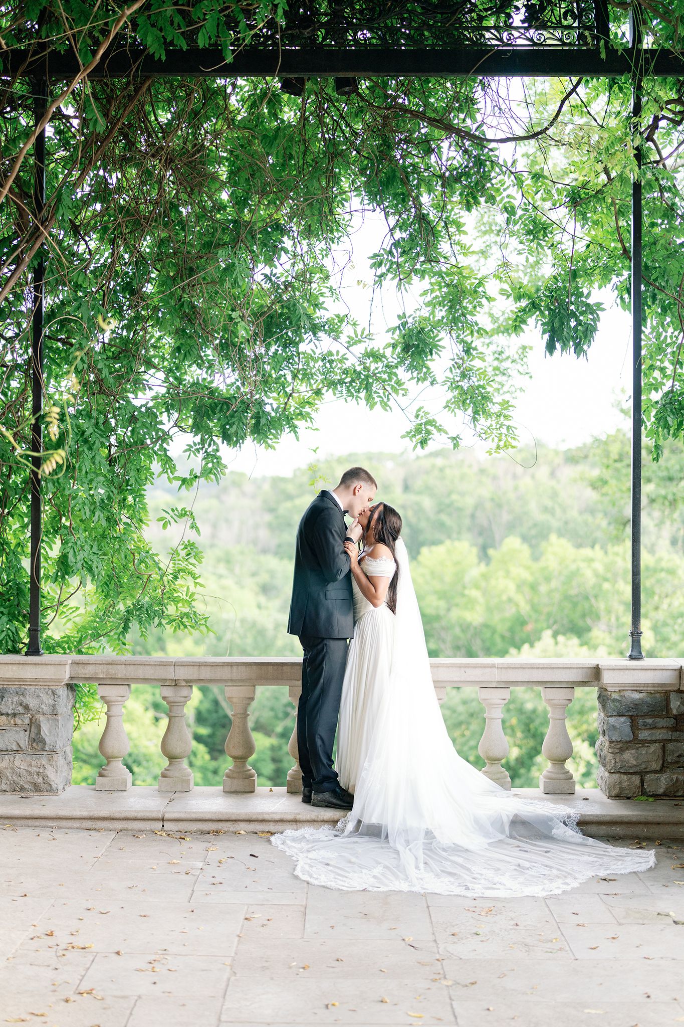 Bride and groom share a romantic kiss under a lush green pergola on an elegant stone balcony.