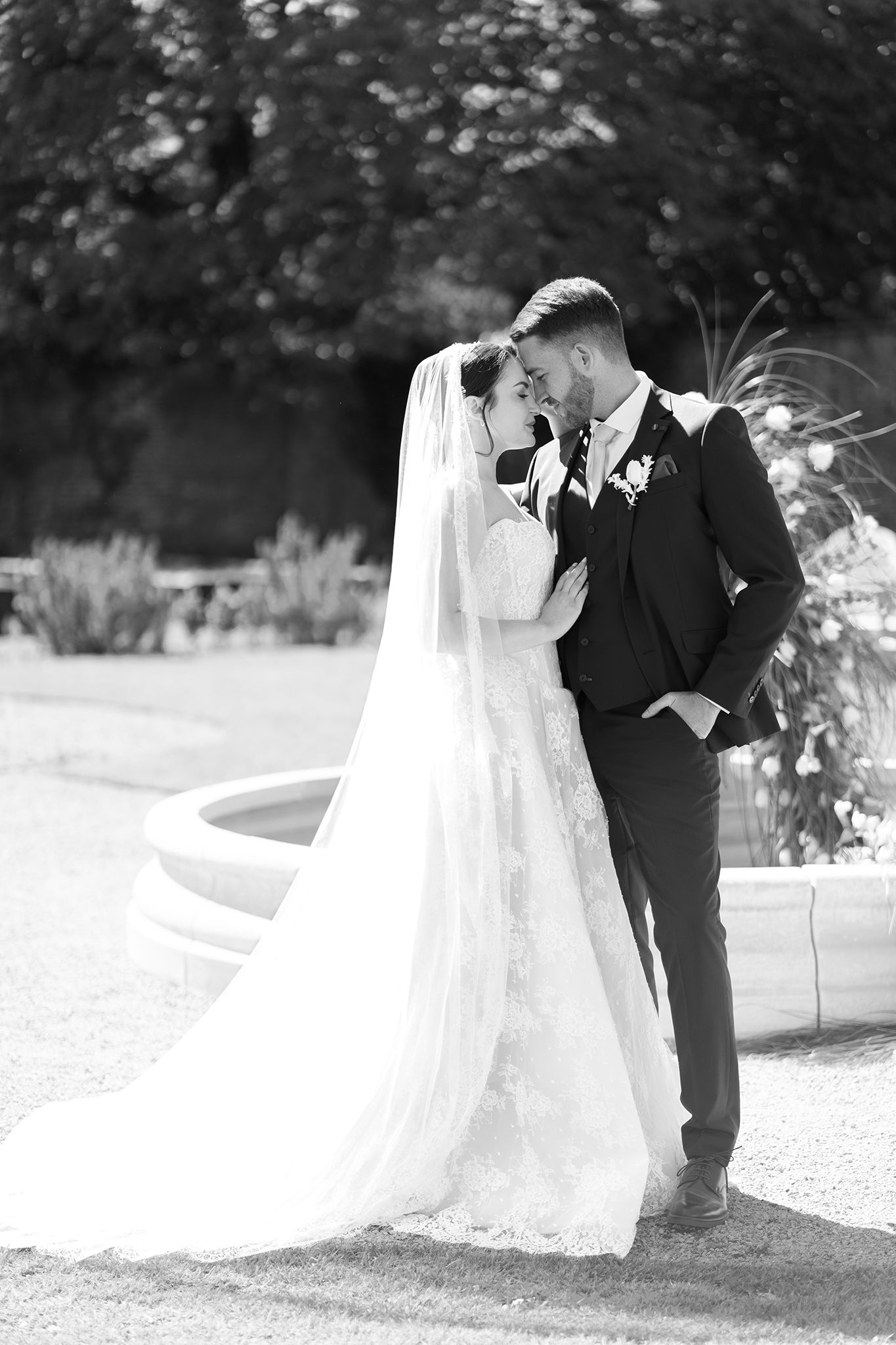 Black and white photo of a bride and groom sharing an intimate moment in a garden, with a fountain in the background.